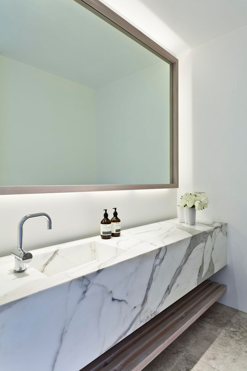 В этой современная ванная комната есть зеркало с подсветкой, мраморный туалетный столик и открытая деревянная полка для дополнительного хранения вещей 