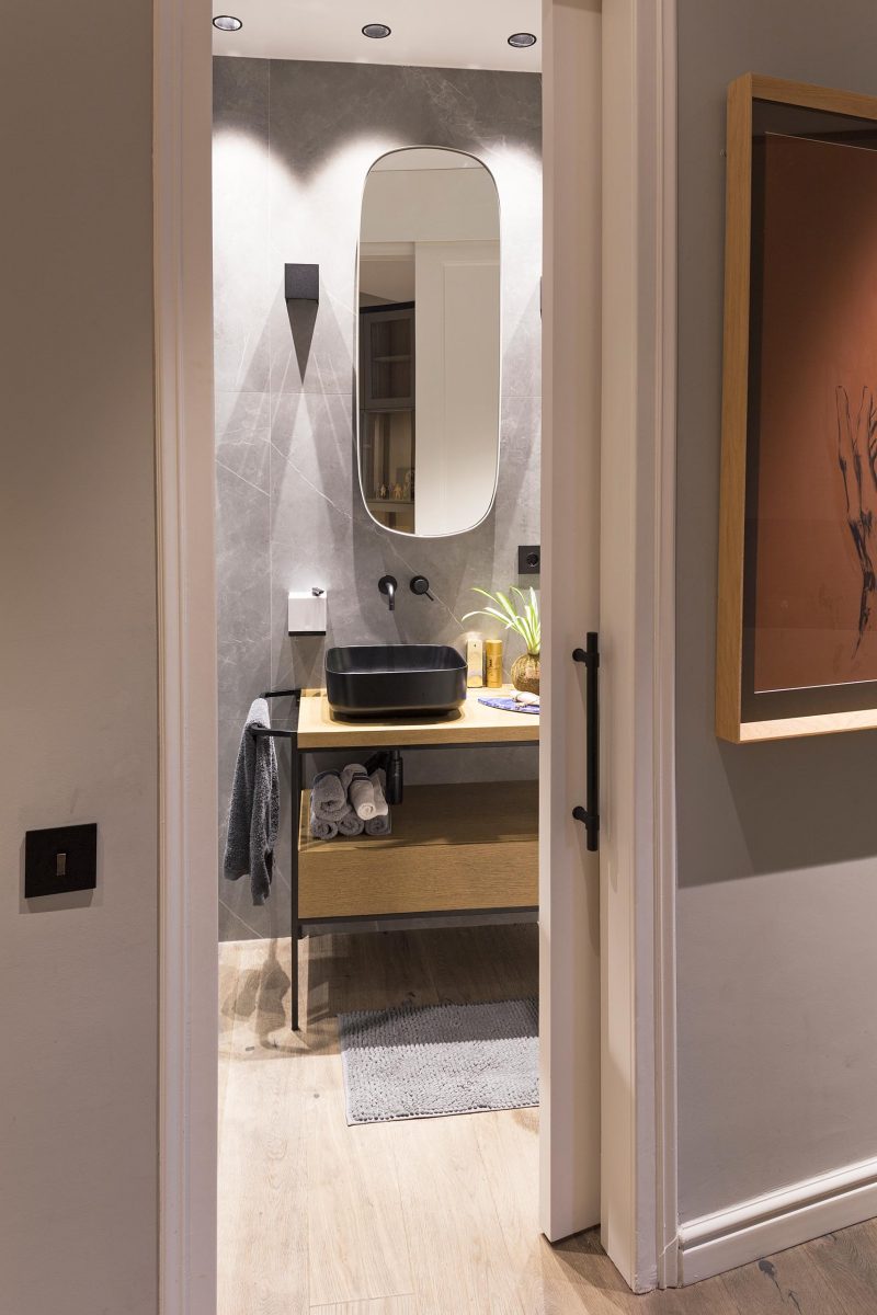 Современная серая ванная комната с зеркалом в форме пилюли, раздвижной дверью, черными вставками и деревянной раковиной.