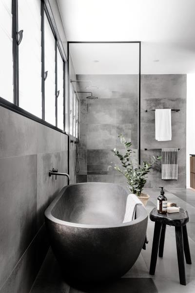 Современная главная ванная комната с серой плиткой большого формата, серая отдельно стоящая ванна и стеклянная душевая перегородка в черной рамке.