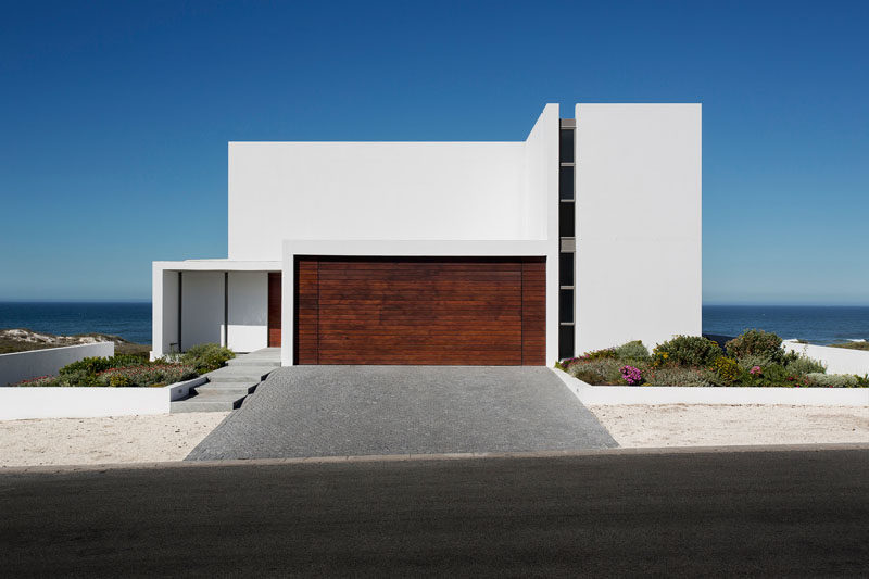 18 вдохновляющих примеров современных гаражных ворот // Гаражные ворота, обшитые панелями из темного дерева, резко контрастируют с полностью белым фасадом и светлым песком вокруг.