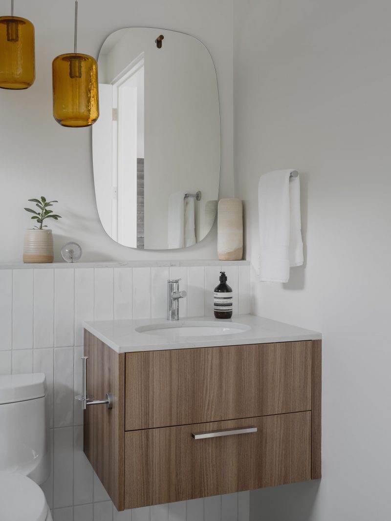 Современная ванная комната с белыми стенами и плиткой, парящей деревянной раковиной и круглым зеркалом.