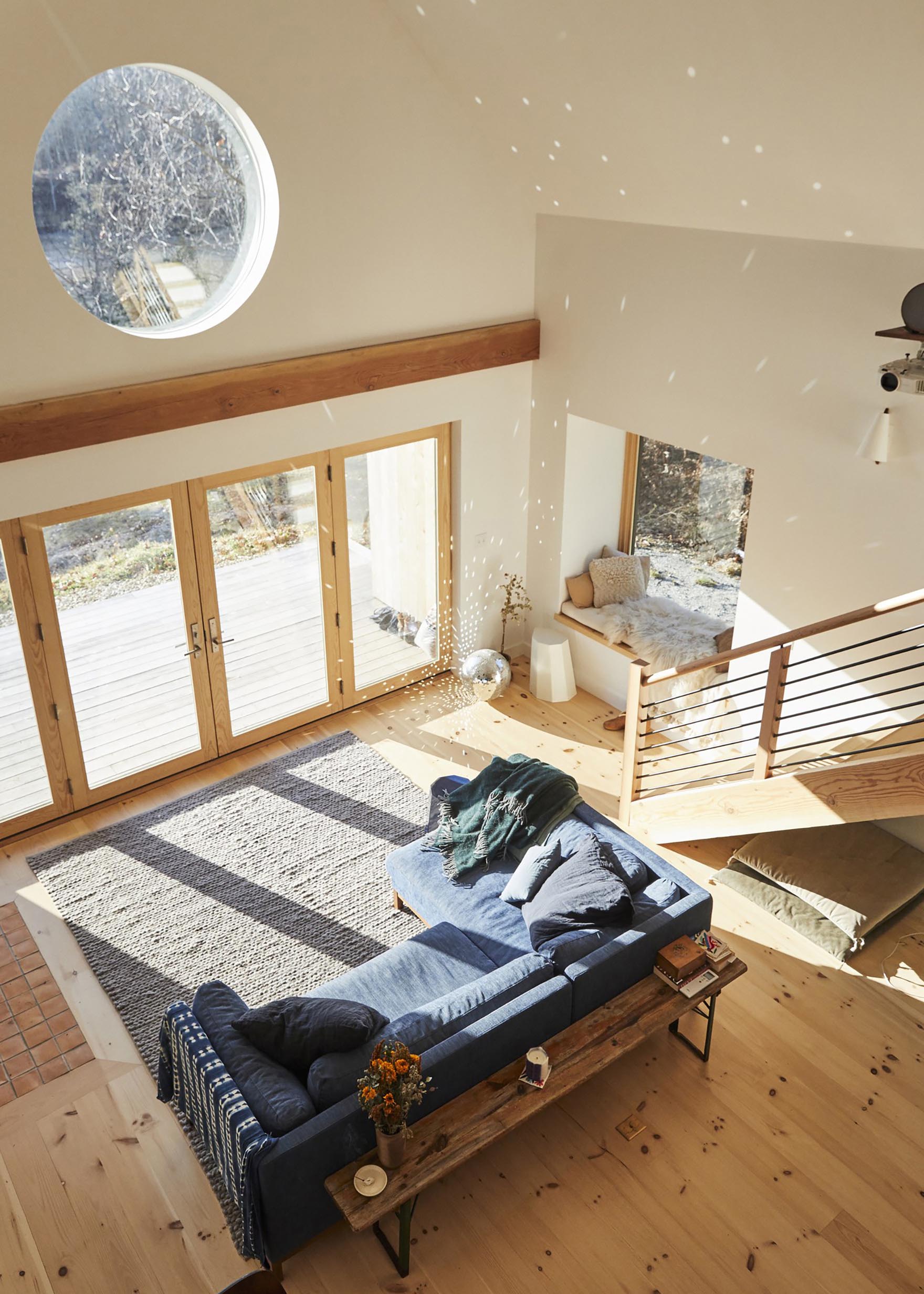 Гостиная в скандинавском стиле с синим диваном, большим серым ковром, уютным сиденьем у окна и зеркальным шаром, рассеивающим солнечный свет.