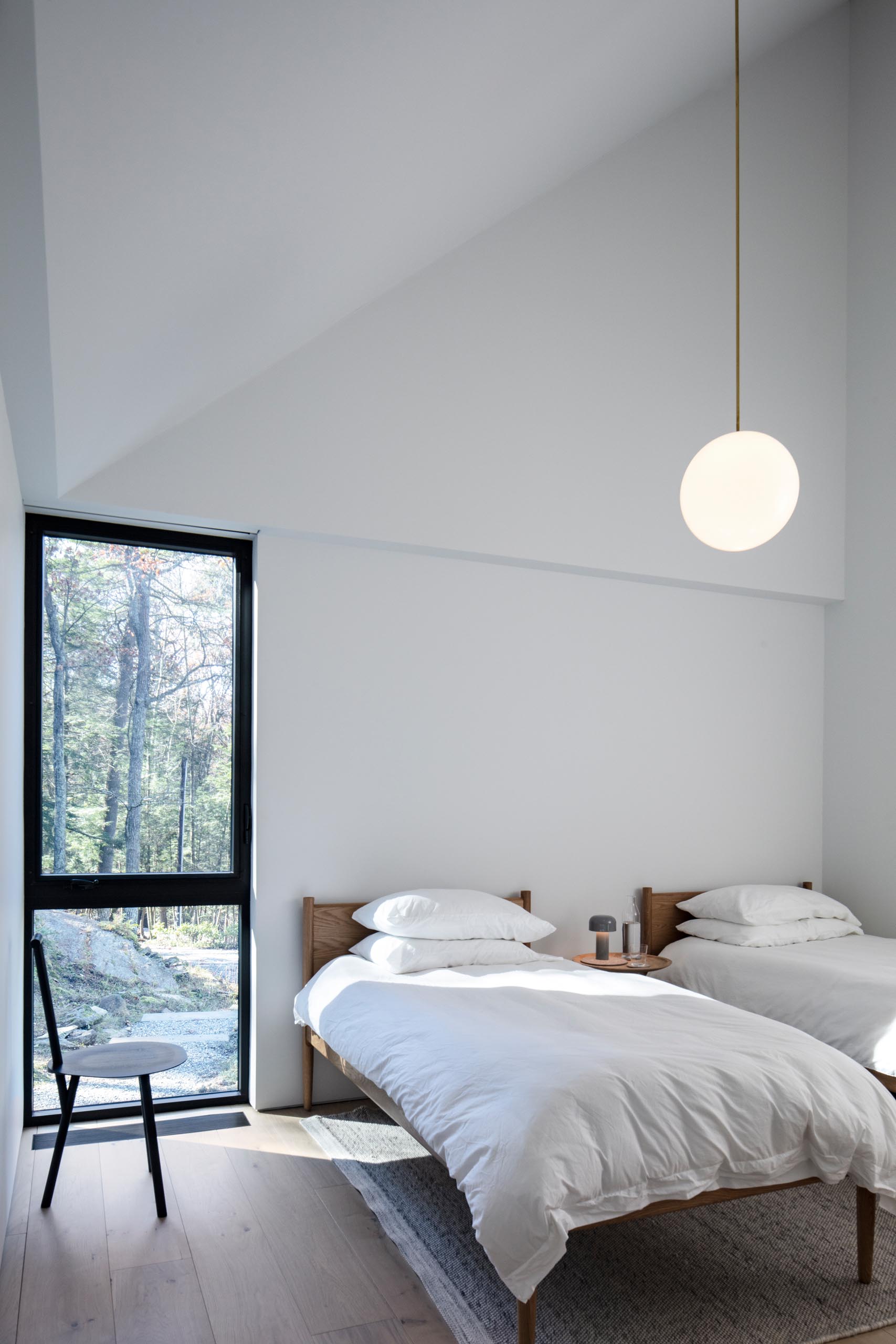Гостевая спальня в стиле минимализма с двумя кроватями, современным черным стулом, белыми стенами и одним подвесным светильником.