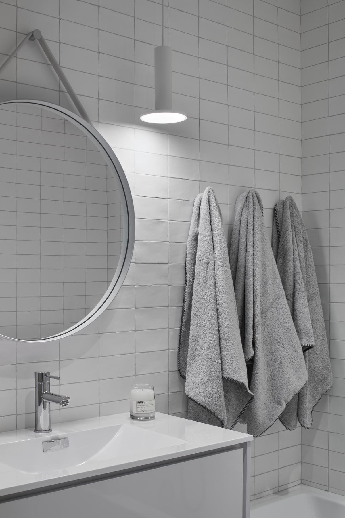 Современная белая ванная комната с белой плиткой, белым подвесным светильником, круглым зеркалом в белой раме и белым туалетным столиком.