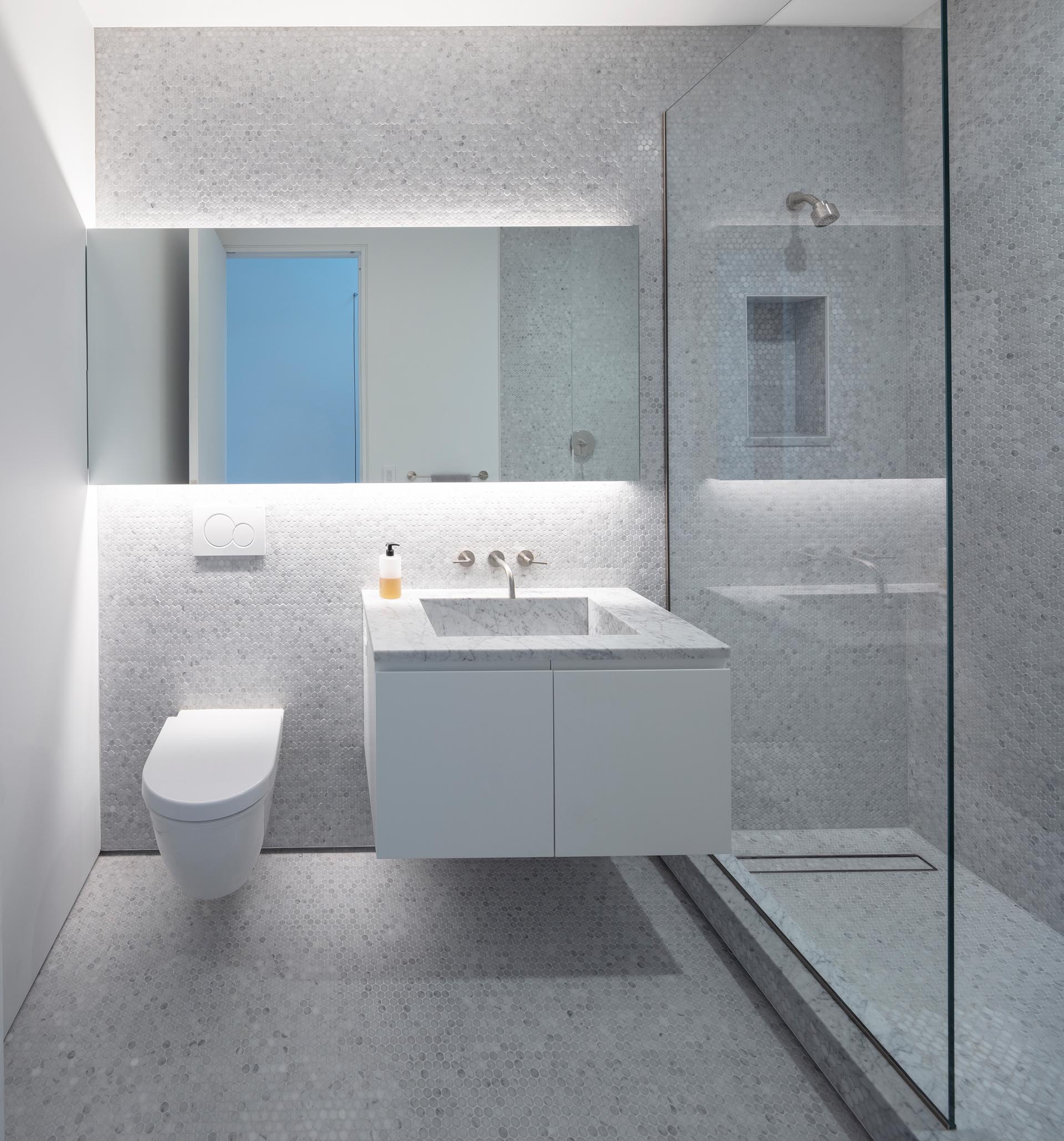 Современная ванная комната с серой плиткой от пола до потолка, стеклянной перегородкой для душа, зеркалом с подсветкой и плавающим туалетным столиком.