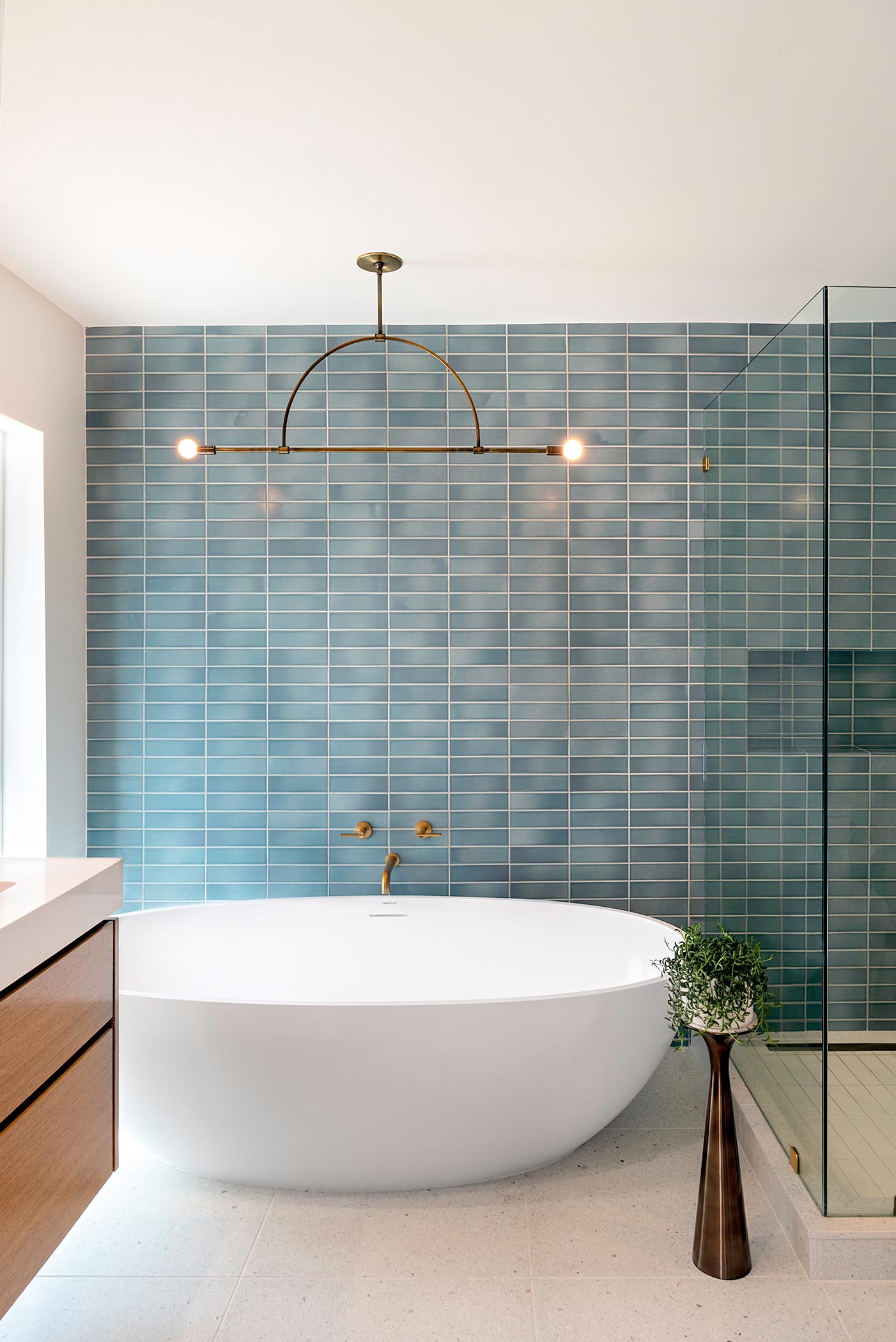 Это современная ванная комната с синей прямоугольной настенной плиткой, минималистичным светильником, отдельно стоящей белой ванной и латунными светильниками.
