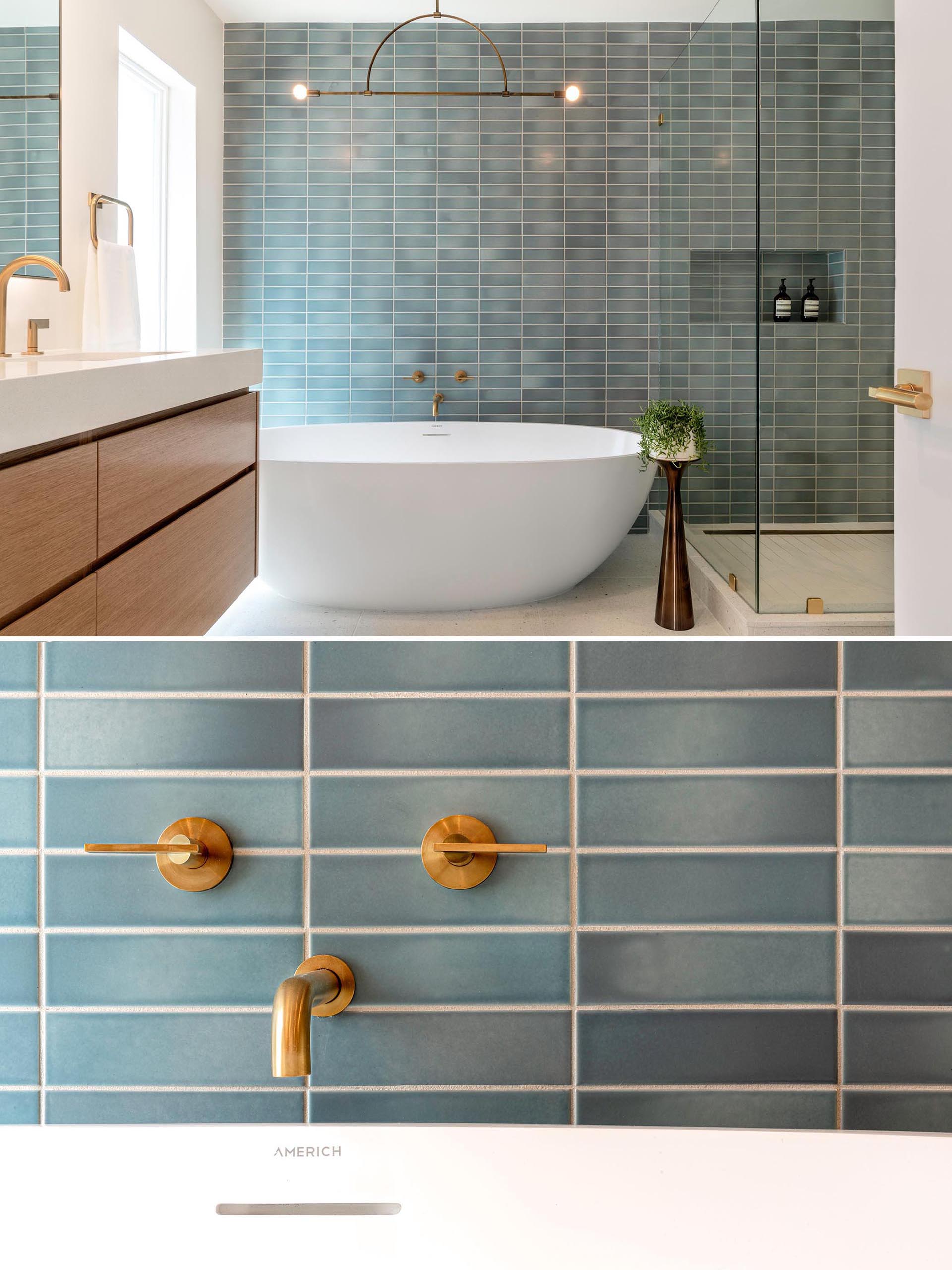 Это современная ванная комната с синей прямоугольной настенной плиткой, минималистичным светильником, отдельно стоящей белой ванной и латунными светильниками.