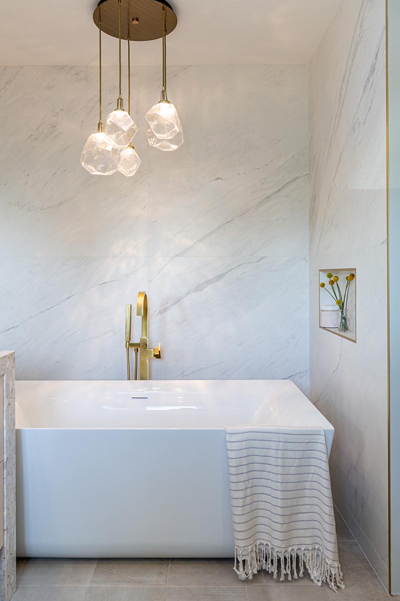 В этой современной ванной комнате есть отдельно стоящая белая ванна, встроенная в нишу. Скульптурный многоликовый подвесной светильник со стеклянными геометрическими абажурами висит над ванной, подчеркивая дизайн. # Ванна # Современная