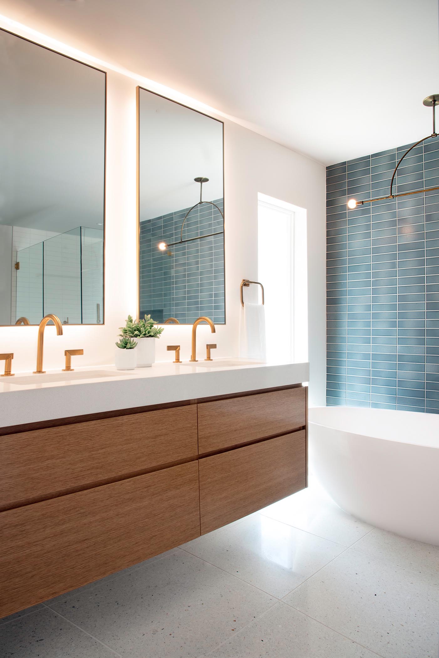 Эта современная ванная комната включает настенный деревянный туалетный столик, сине-белую прямоугольную настенную плитку, минималистичный светильник, зеркала с подсветкой, отдельно стоящую белую ванну и латунные светильники.