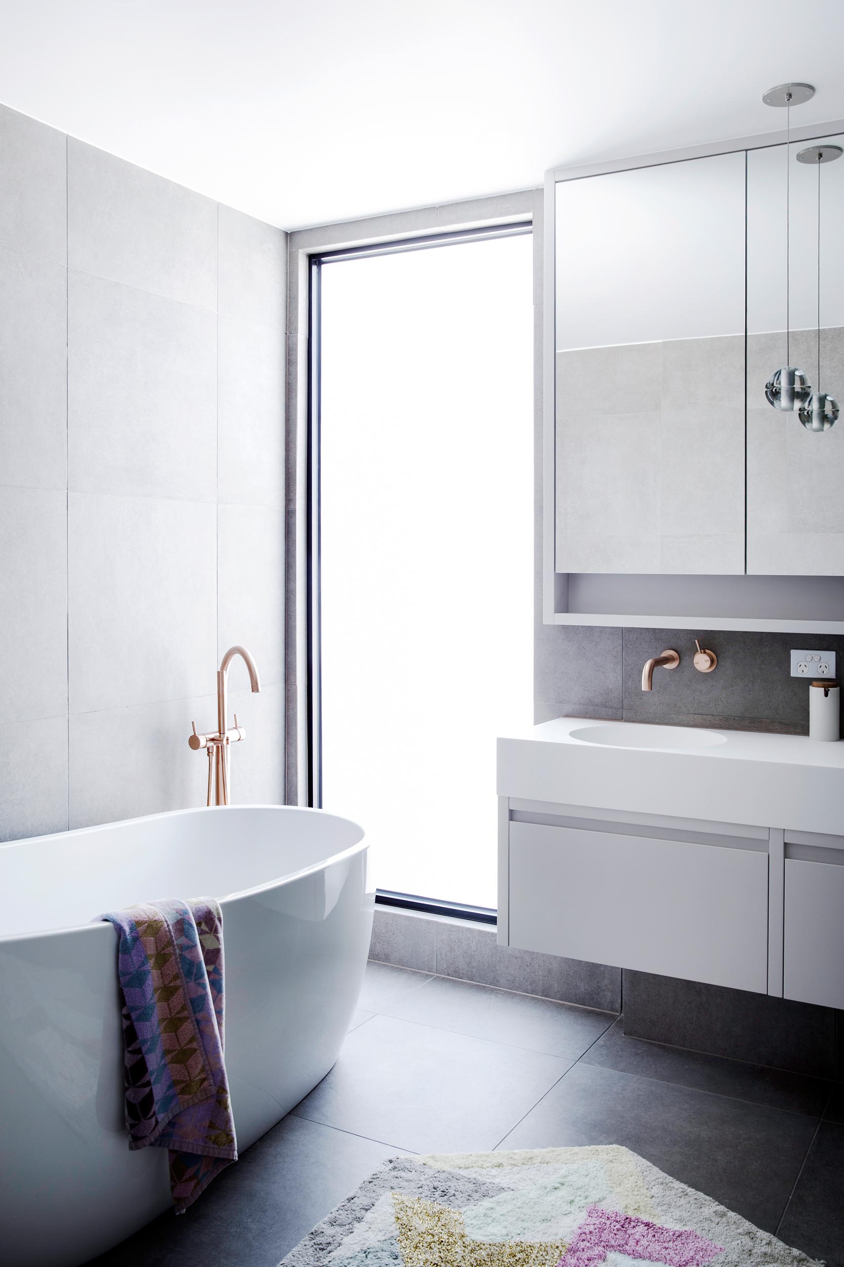 Современная ванная комната с большим вертикальным окном, через которое отдельно стоящая ванна наполняется естественным светом.