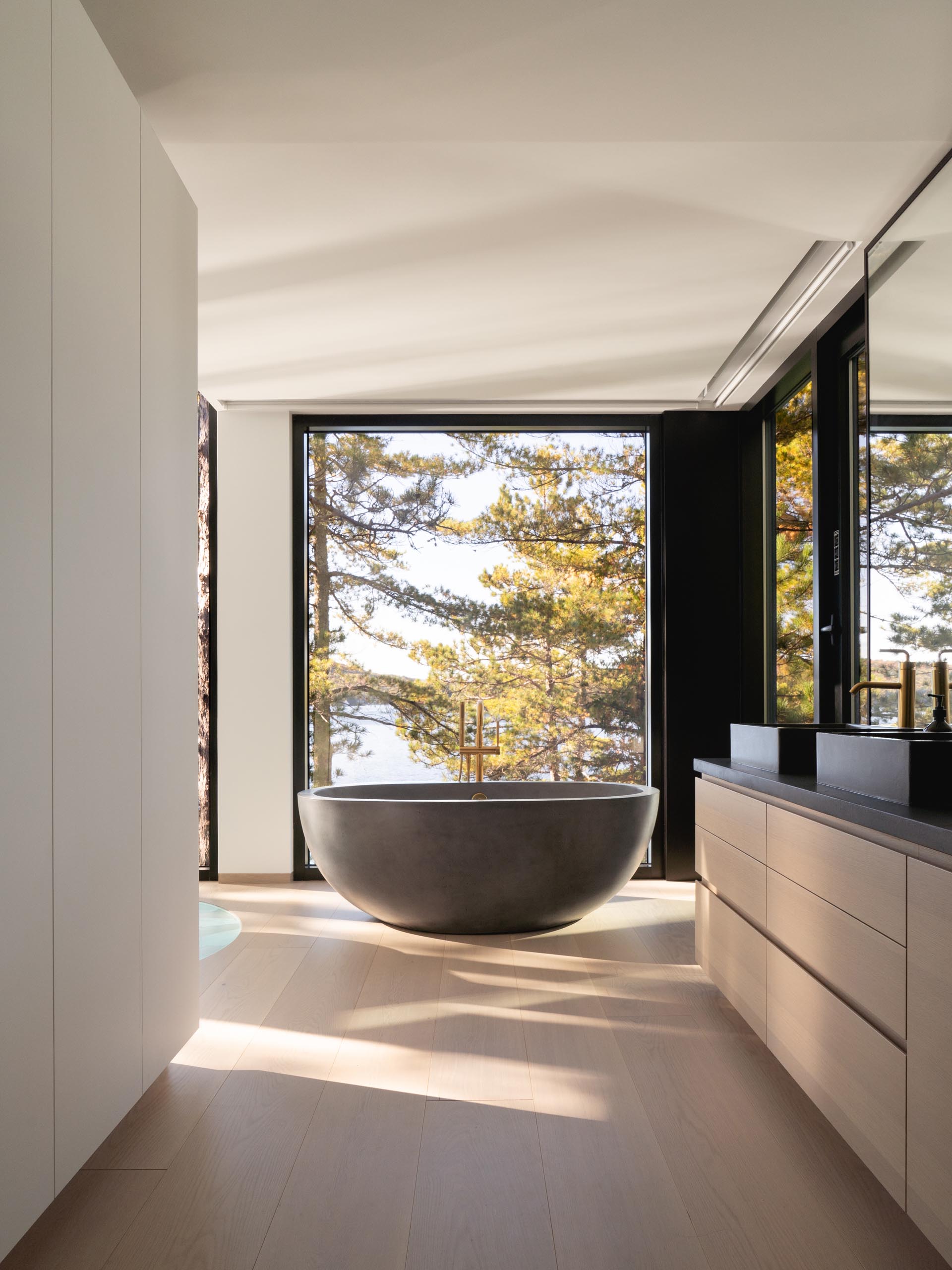 Современная ванная комната с отдельно стоящей ванной перед окном и минималистский деревянный туалетный столик с темной столешницей.