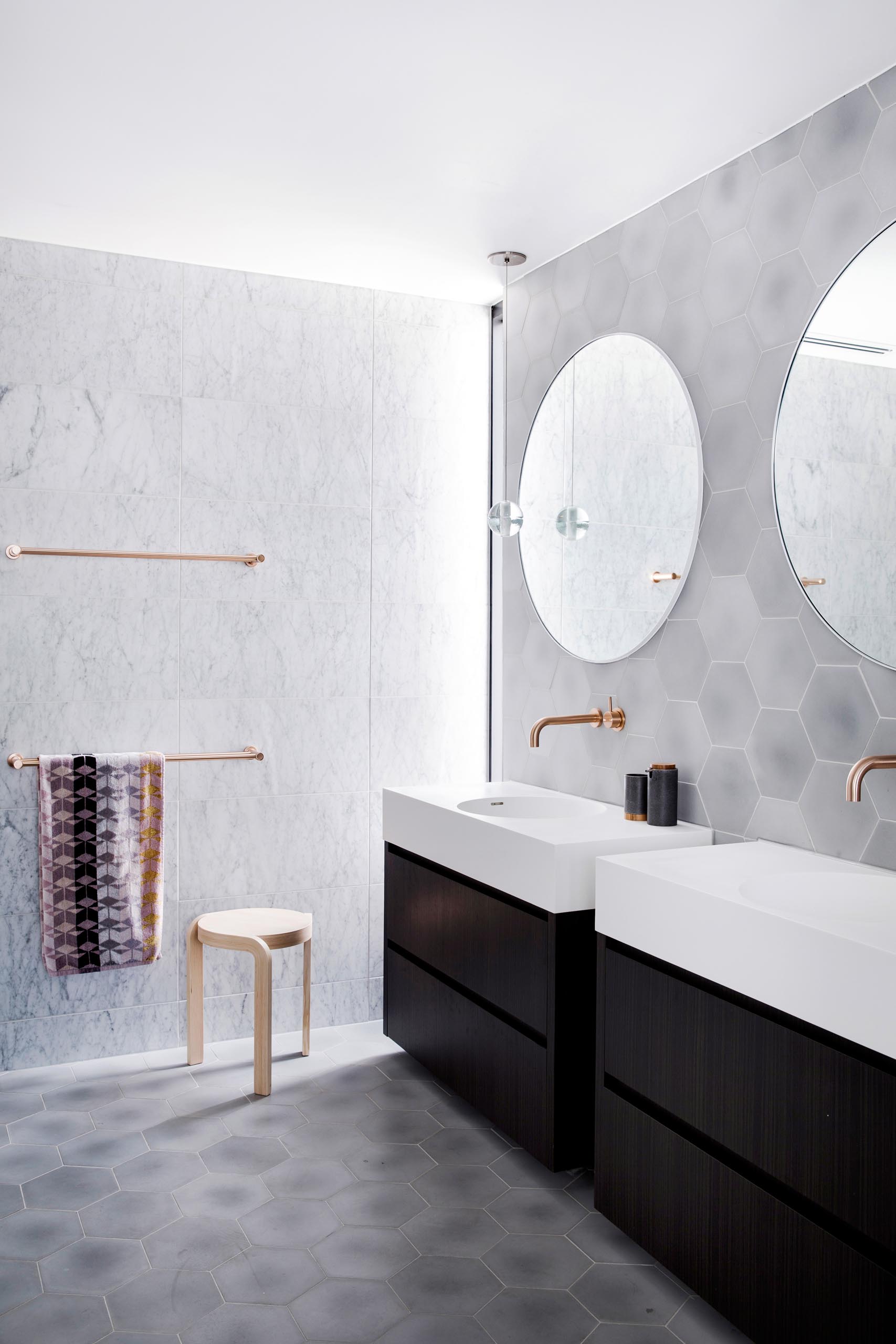 Входя в эту главную ванную комнату, естественный свет из высокого узкого окна в углу освещает вешалки для полотенец из полированного розового золота, прикрепленные к стене, покрытые плиткой большого формата.