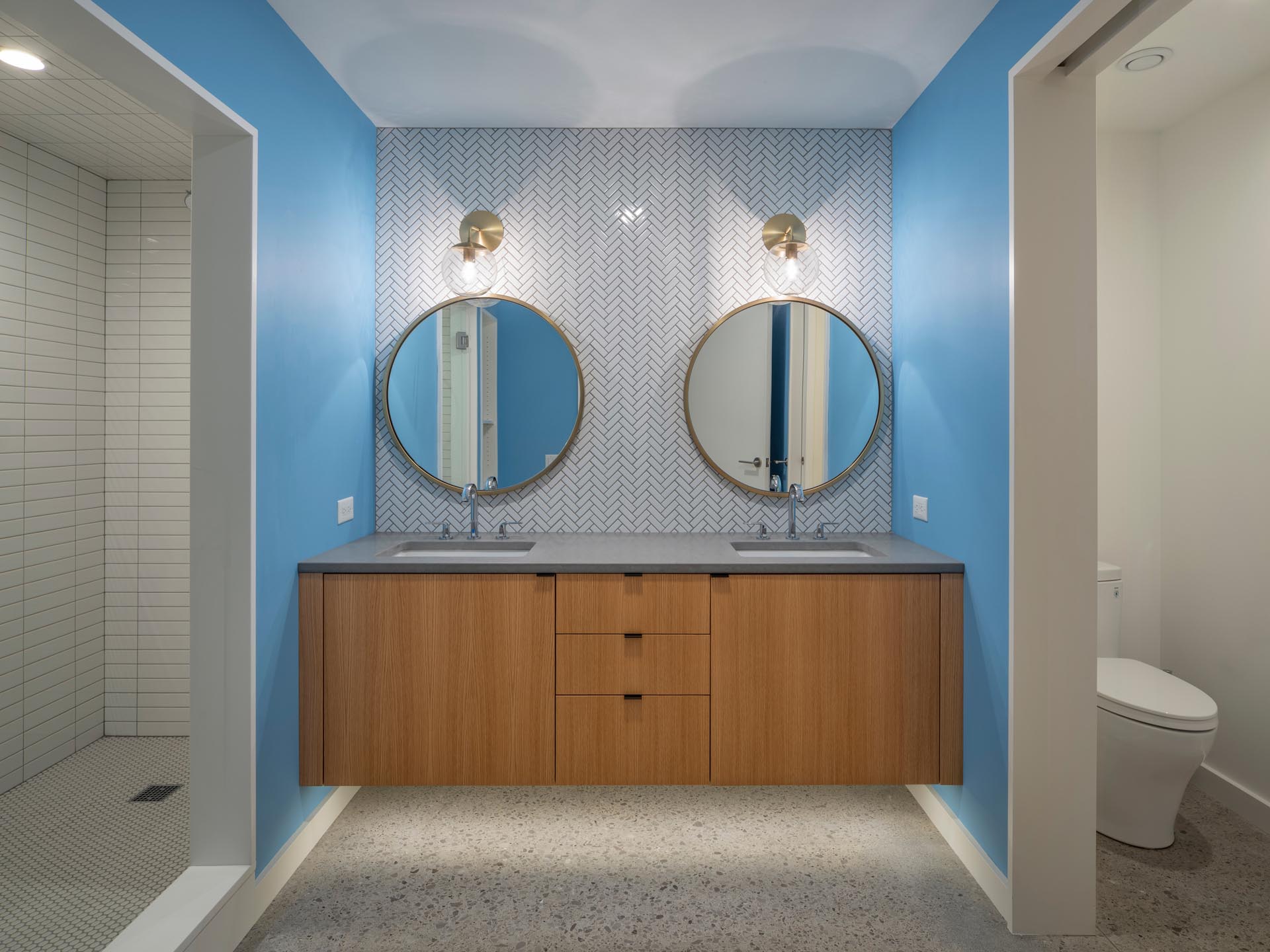 Современная ванная комната с ярко-синими стенами, которые добавляют яркости, белый - парящий деревянный туалетный столик с подсветкой под ним, а наверху - два круглых зеркала с бронзовыми рамами, которые сочетаются с бра.