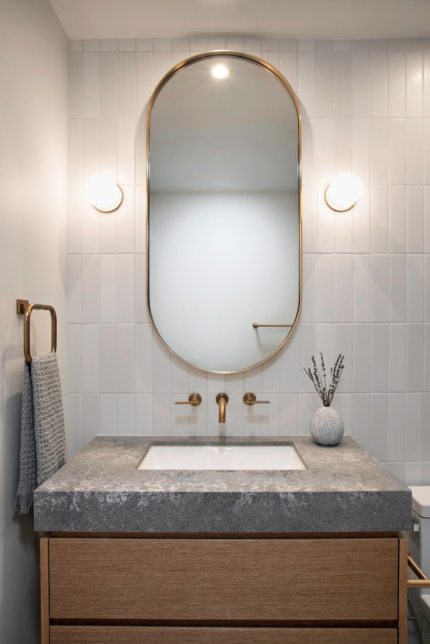 В этой ванной комнате есть нестандартное зеркало в форме пилюли, окруженное специальными настенными бра, которое висит над деревянным туалетным столиком с серой столешницей и белой раковиной под столешницей.