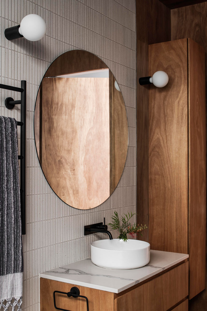 Эта современная ванная комната имеет облицованные плиткой стены, деревянный туалетный столик и подходящую мебель, а также круглое зеркало с бра с обеих сторон. # Современная ванная # Круглое зеркало # Деревянная тщета # Бра # Плиточная стена