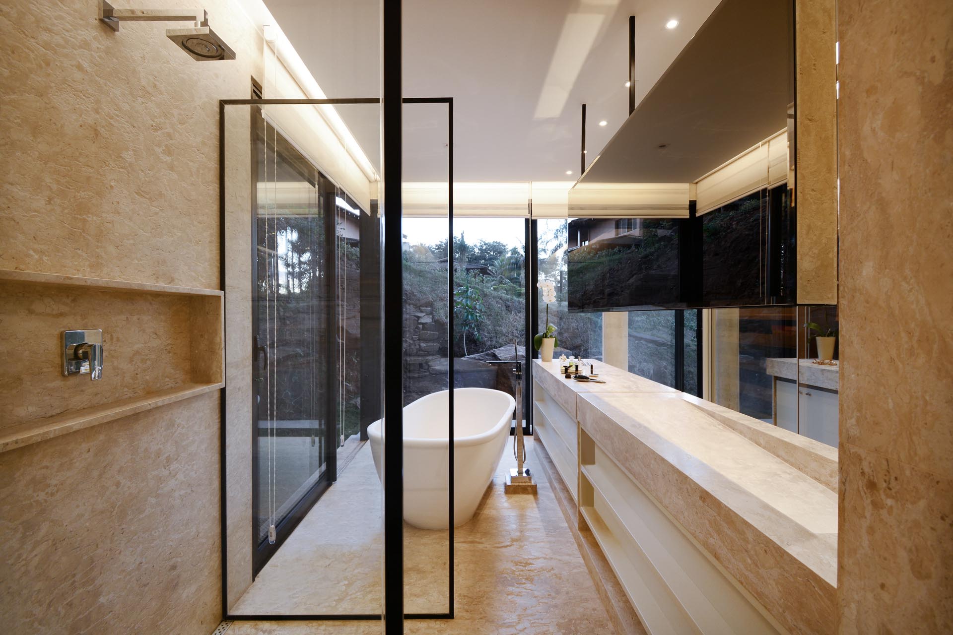 Современная ванная комната с окнами от пола до потолка, из которых открывается вид на деревья из отдельно стоящей ванны.
