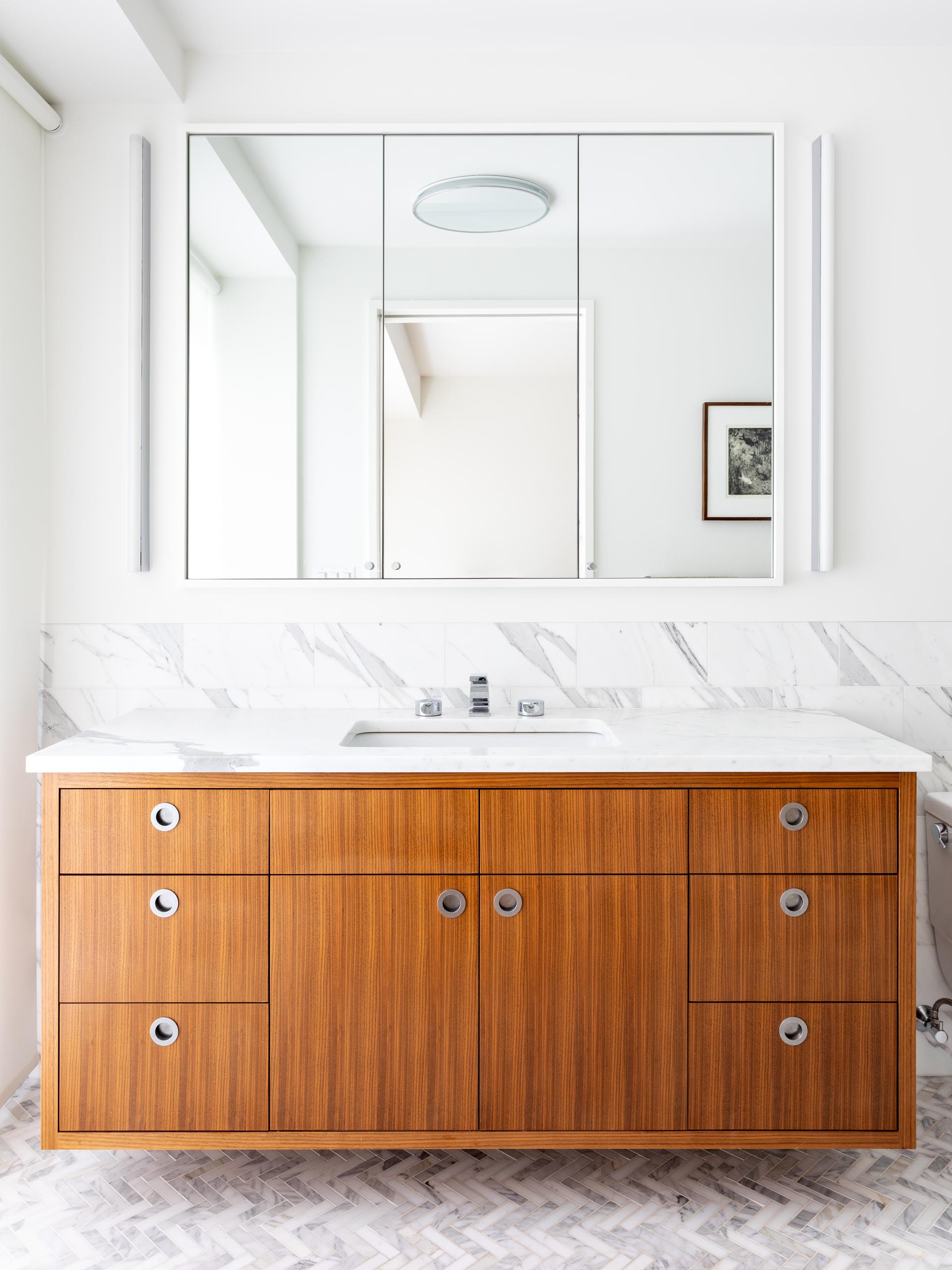 В этой современной ванной комнате есть парящий деревянный туалетный столик с трехпанельным зеркалом, прикрепленным к стене, и мраморный фартук, который продолжается до душа.