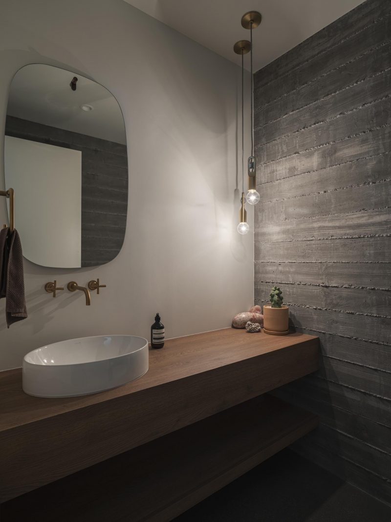 Современная ванная комната с бетонной стеной из досок, деревянной раковиной и круглым зеркалом.