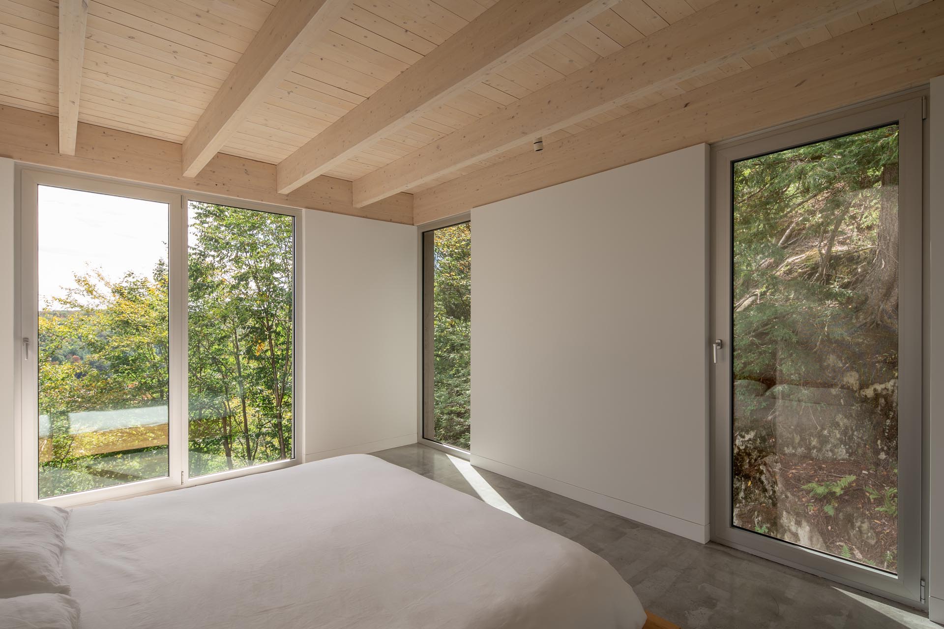 В главной спальне с минималистичной мебелью есть высокие окна, обрамляющие деревья и скалы снаружи.