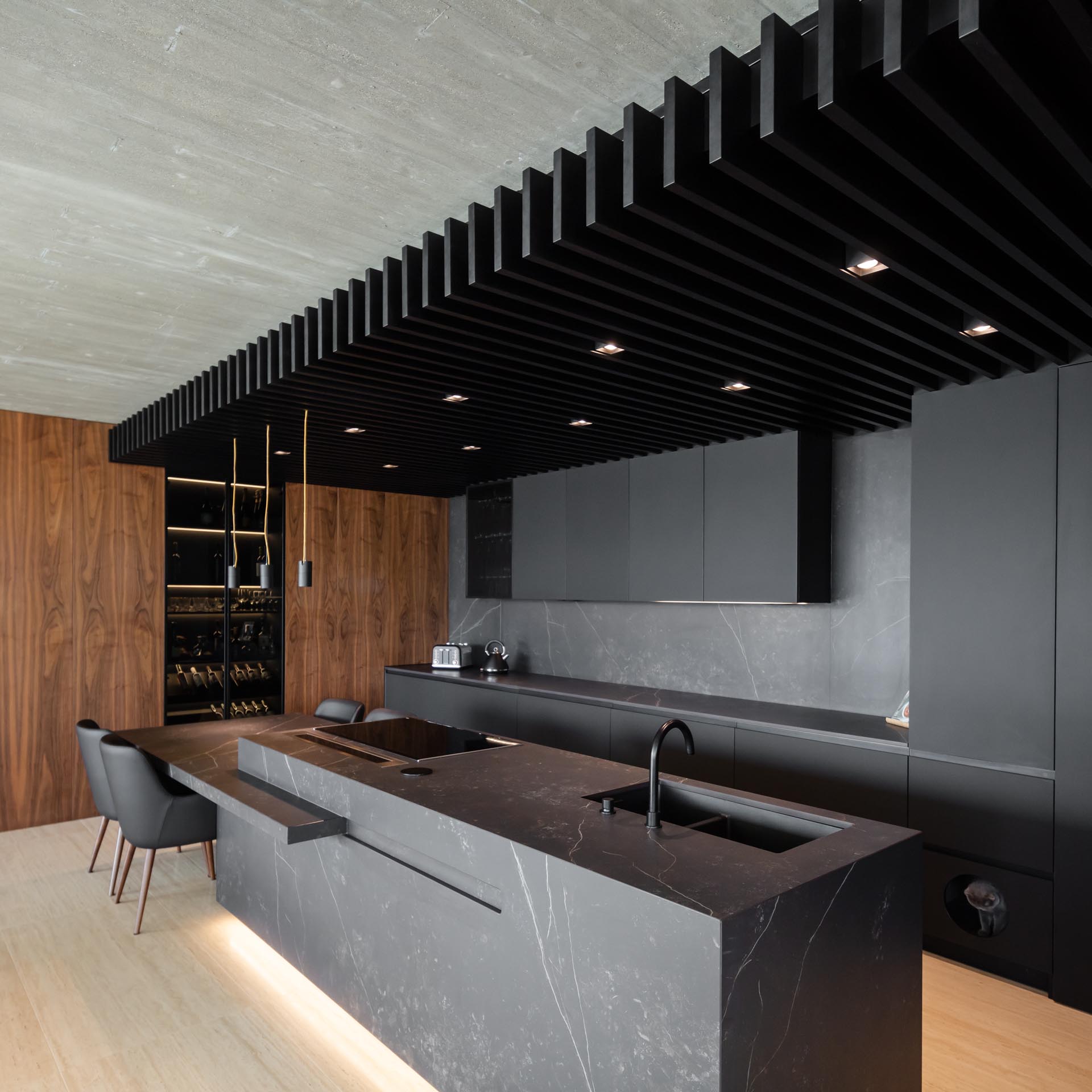 Современная кухня со стеной из матовых черных минималистичных шкафов (в которой также есть небольшая дверца для доступа к ящику для кошачьих туалетов), а также потолок из черного дерева с равномерно распределенным освещением между планками.