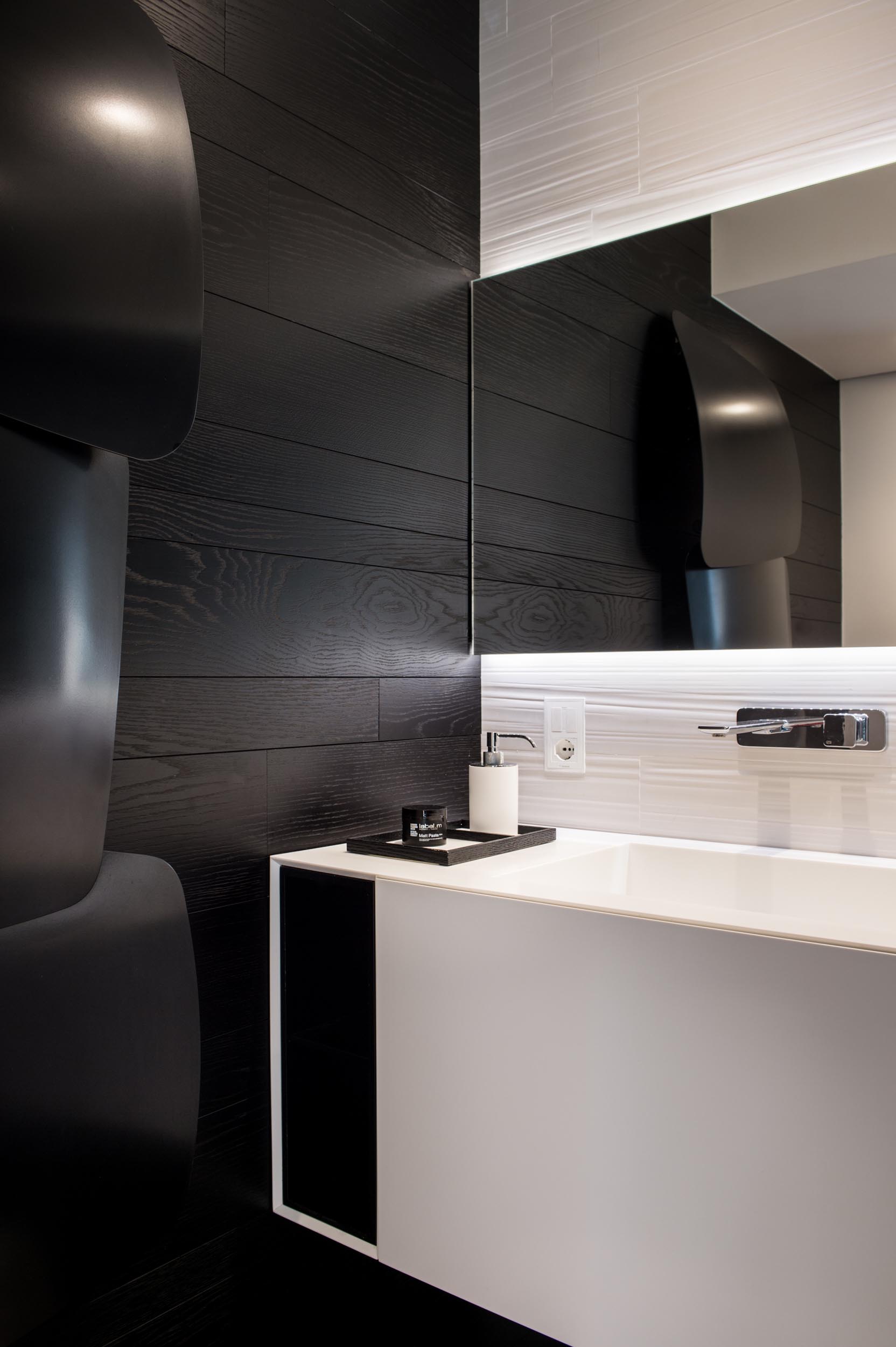 Современная ванная комната с белым туалетным столиком и зеркалом с подсветкой, а также контрастной черной акцентной стеной.