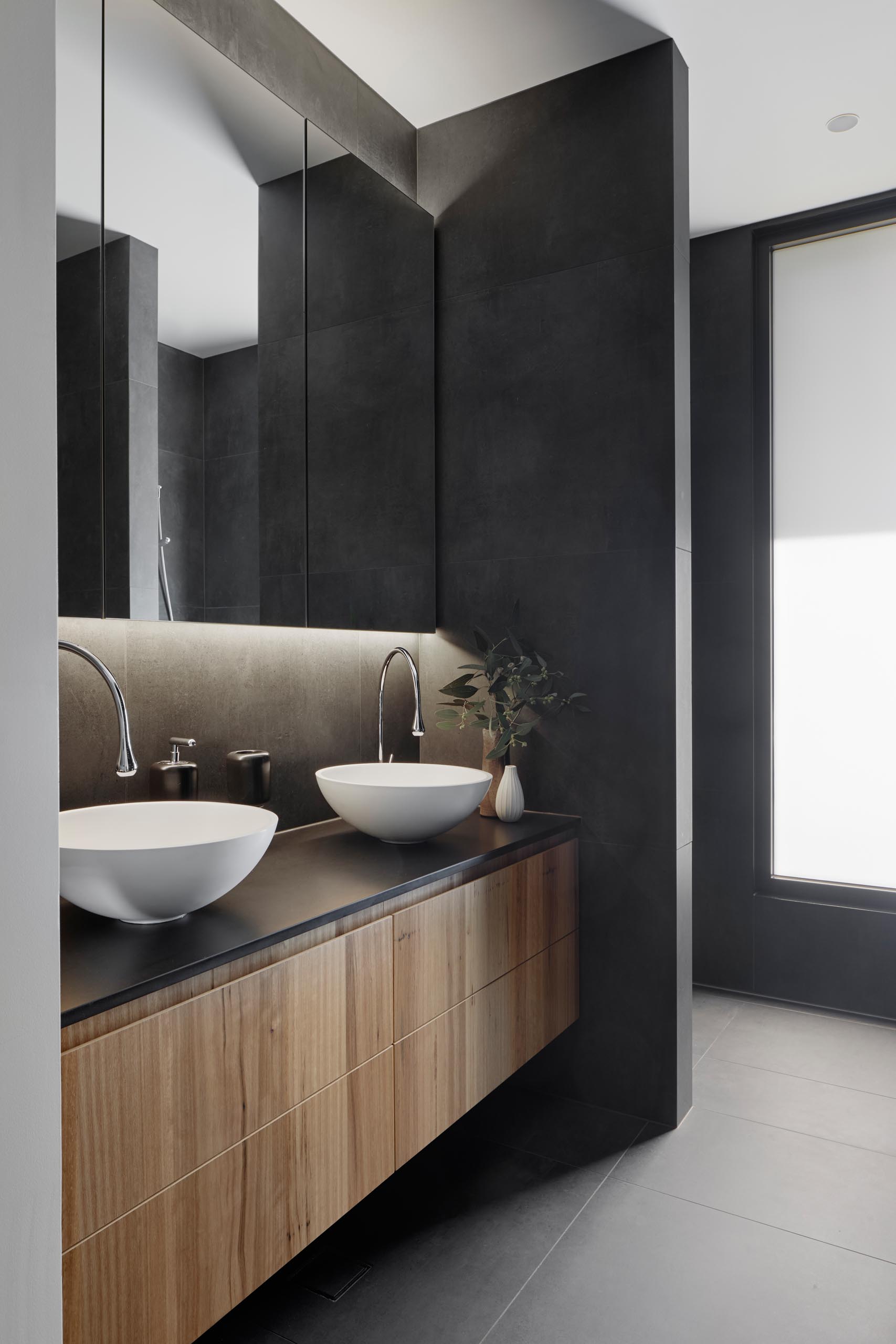 В этой современной ванной комнате темно-серые стены и пол сочетаются с деревянной раковиной, белыми раковинами и зеркалом с подсветкой.