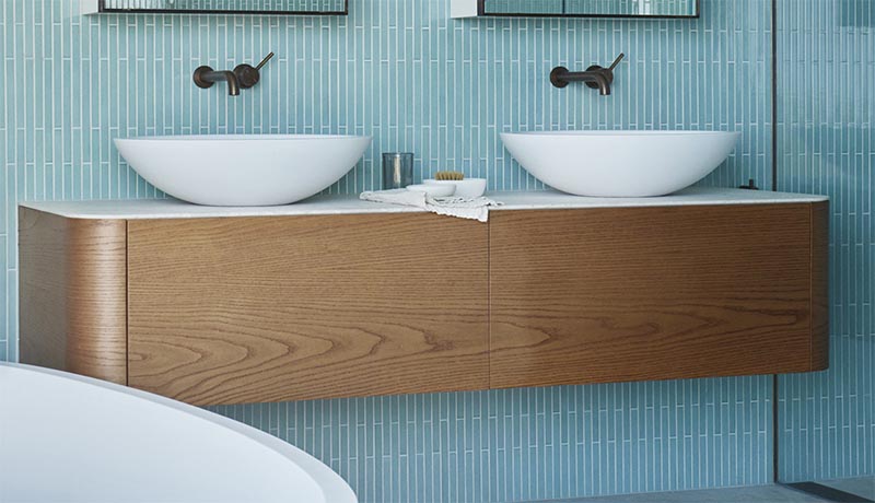 В этой современной ванной комнате есть парящий деревянный туалетный столик с изогнутыми углами, добавляющий мягкости пространству, а на тонкой белой столешнице находится пара минималистичных белых раковин. #FloatingVanity #WoodVanity # СовременныеВанная