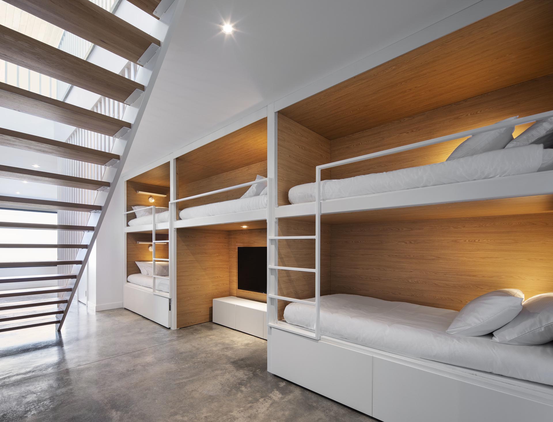У подножия этой современной лестницы, ведущей в коридор, стоят несколько комплектов двухъярусных кроватей. Эти двухъярусные кровати имеют интерьер из дерева, с белыми ужинами и лестницами, которые сочетаются со стенами.