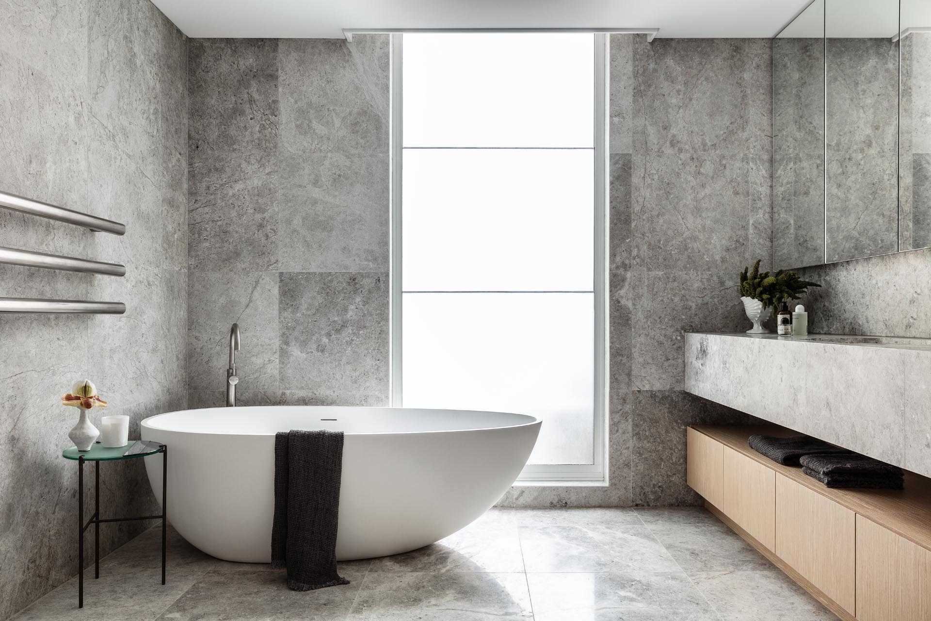 В этой современной главной ванной комнате натуральный камень с серой отделкой покрывает стены и пол, а также туалетный столик. Матовое окно от пола до потолка пропускает свет, но при этом сохраняет уединение.