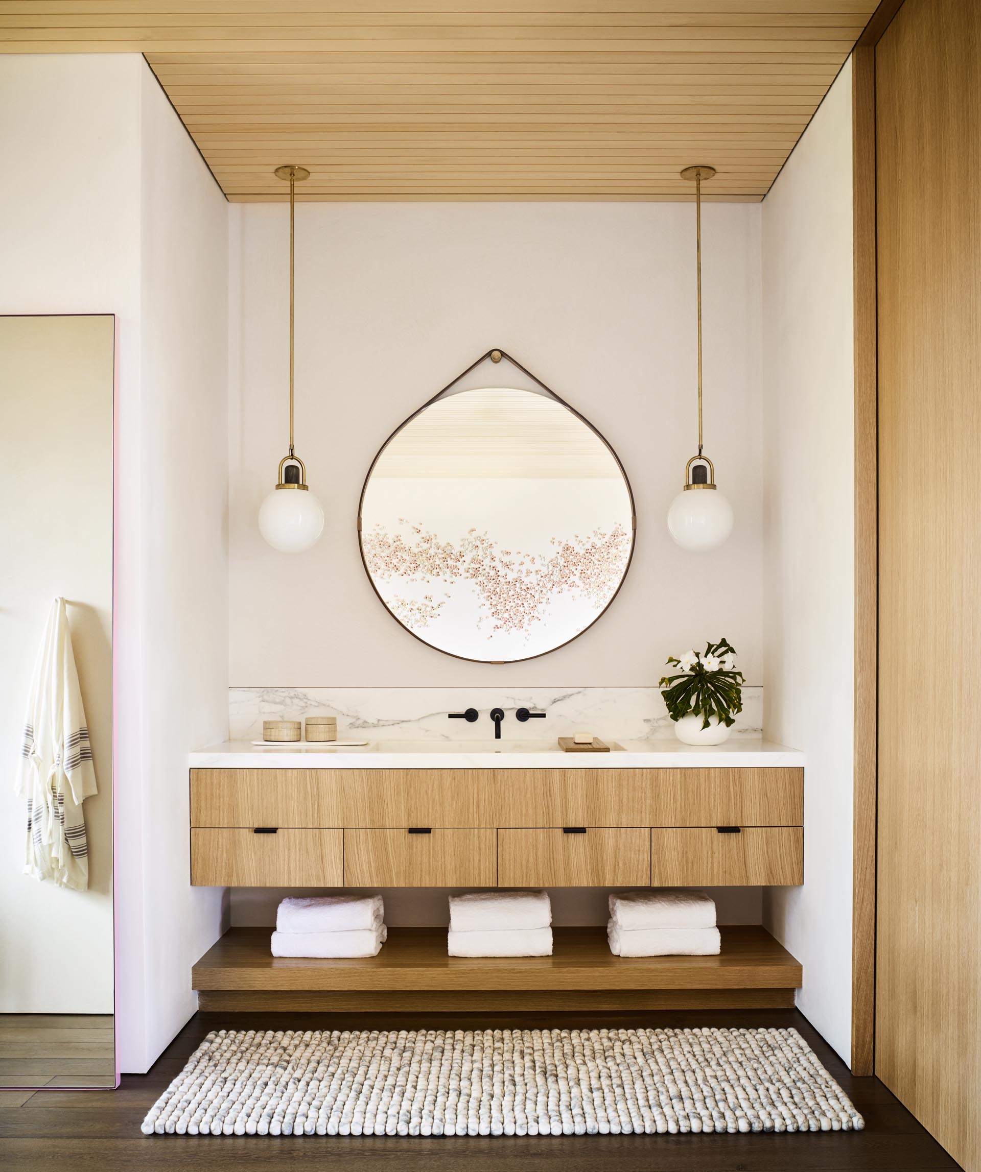 Современная ванная комната с естественной цветовой палитрой, туалетным столиком из светлого дерева и круглым подвесным зеркалом.