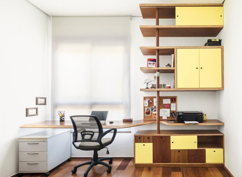 В этом современном домашнем офисе есть специально разработанный стол, который заполняет ширину стены, и деревянный стеллаж с желтыми дверцами, который добавляет мягкий оттенок цвета. # Домашний офис # Современный стол # Стеллажи