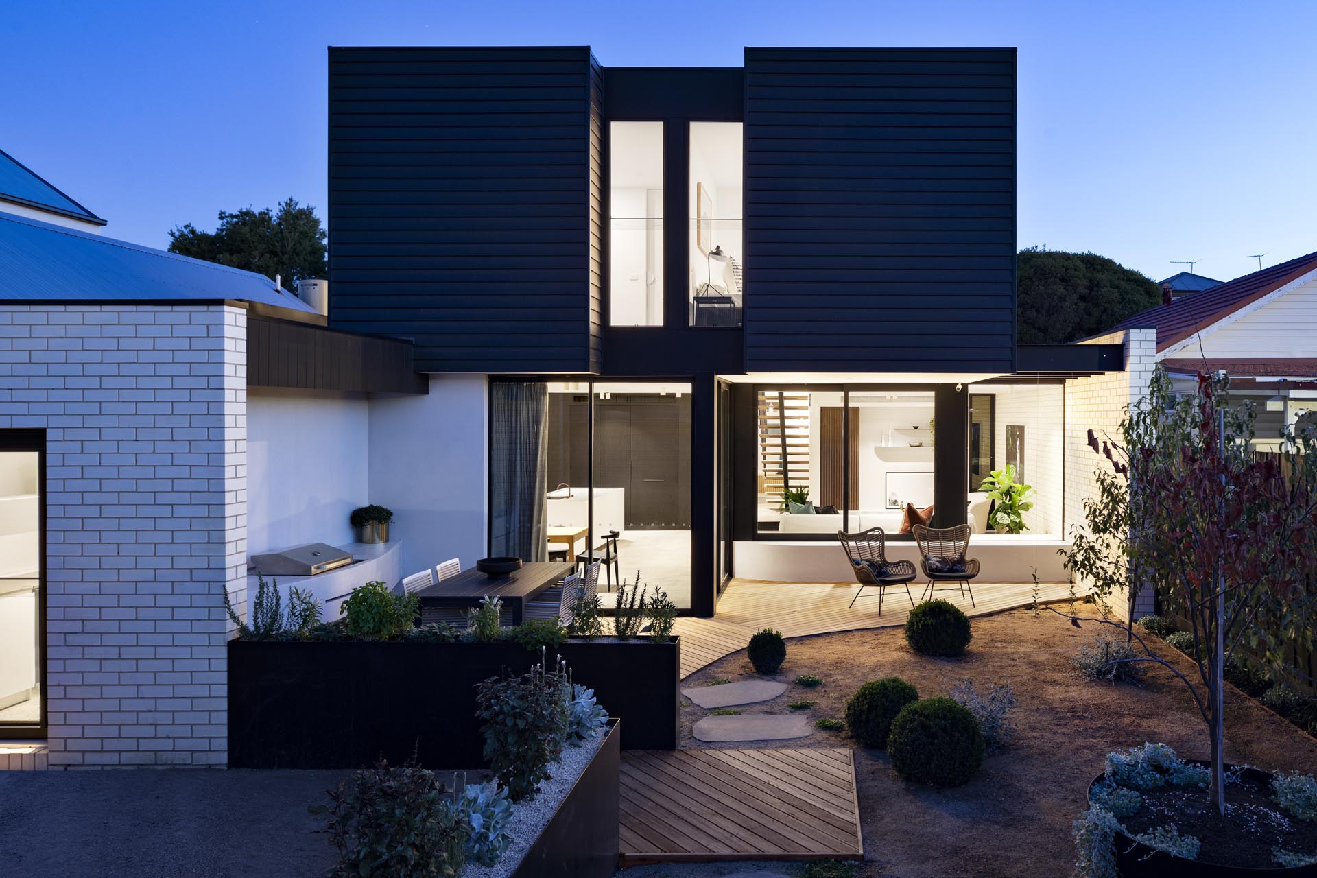 Современная пристройка к дому с черным фасадом, благоустроенным двором с барбекю, обеденной зоной на открытом воздухе и патио.