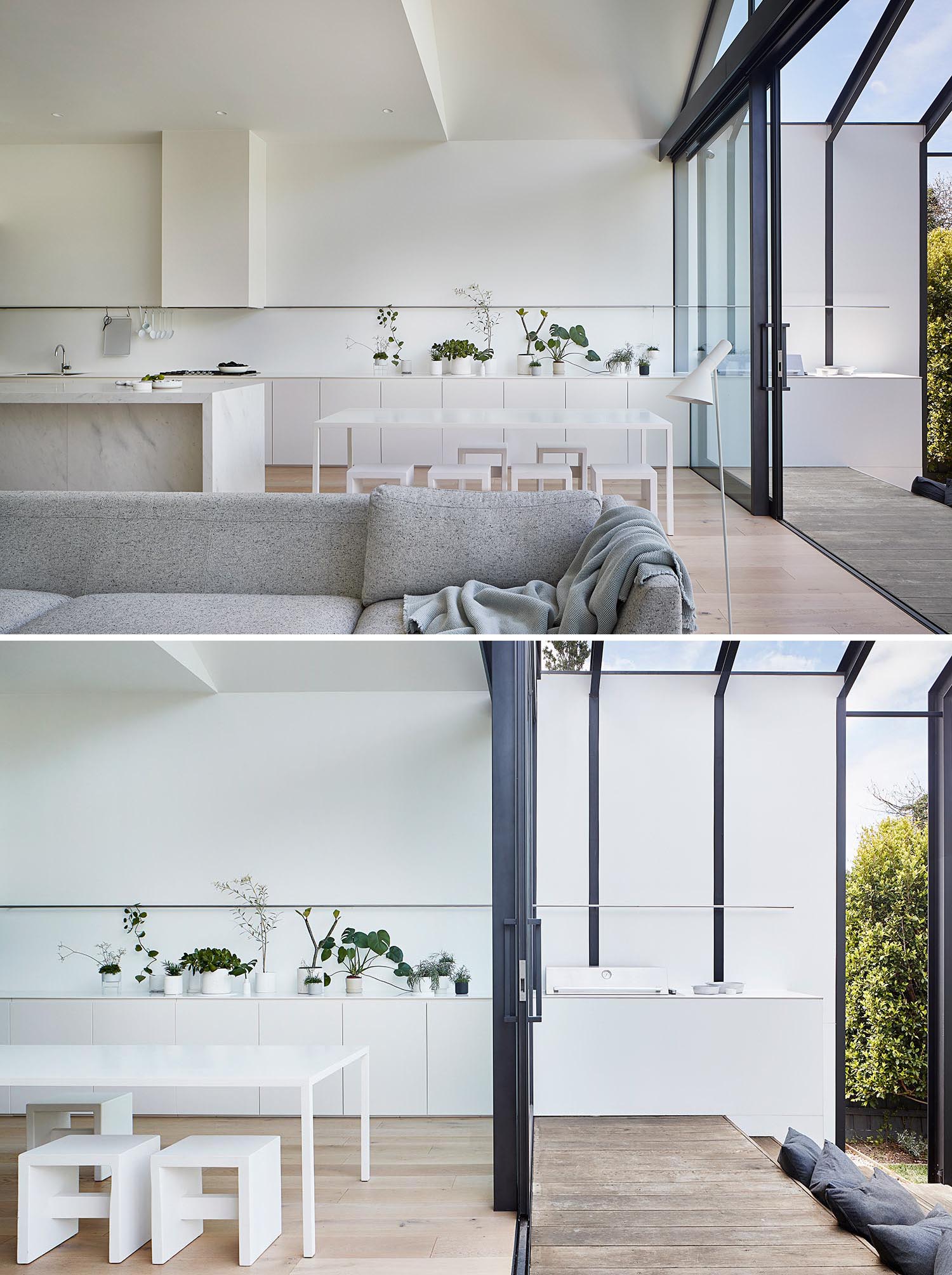 Современная пристройка к дому с белой кухней, большой раздвижной стеклянной дверью и зоной для барбекю на открытом воздухе.