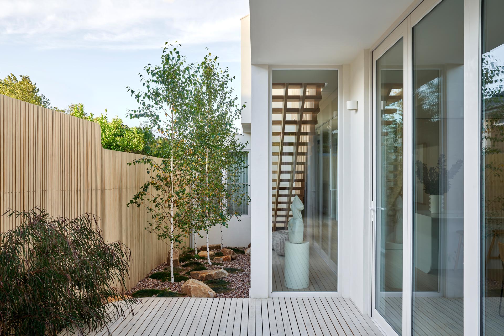 Современный дом с боковым садом с забором из светлого дерева, камнями, травой и деревьями.