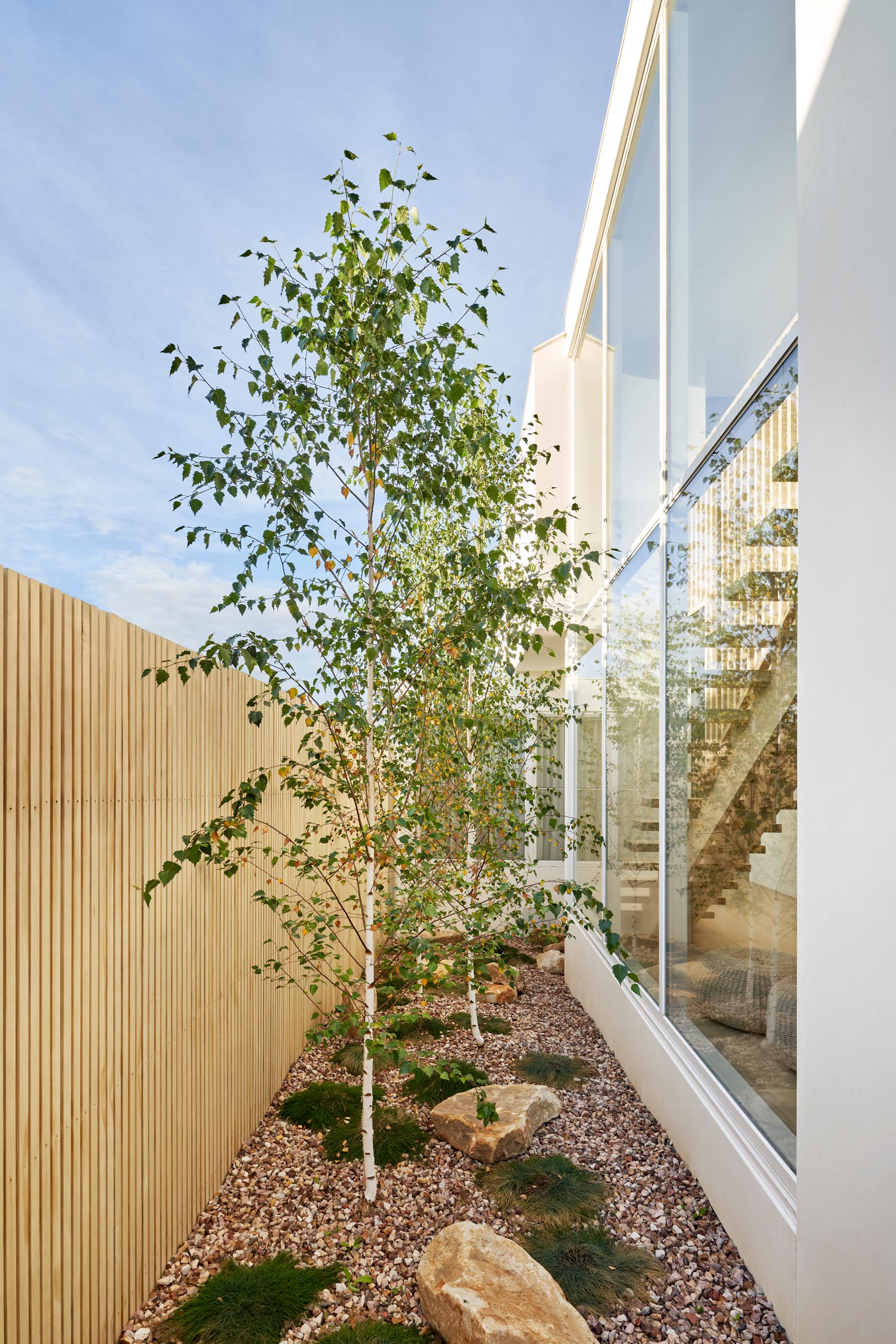 Современный дом с боковым садом с забором из светлого дерева, камнями, травой и деревьями.