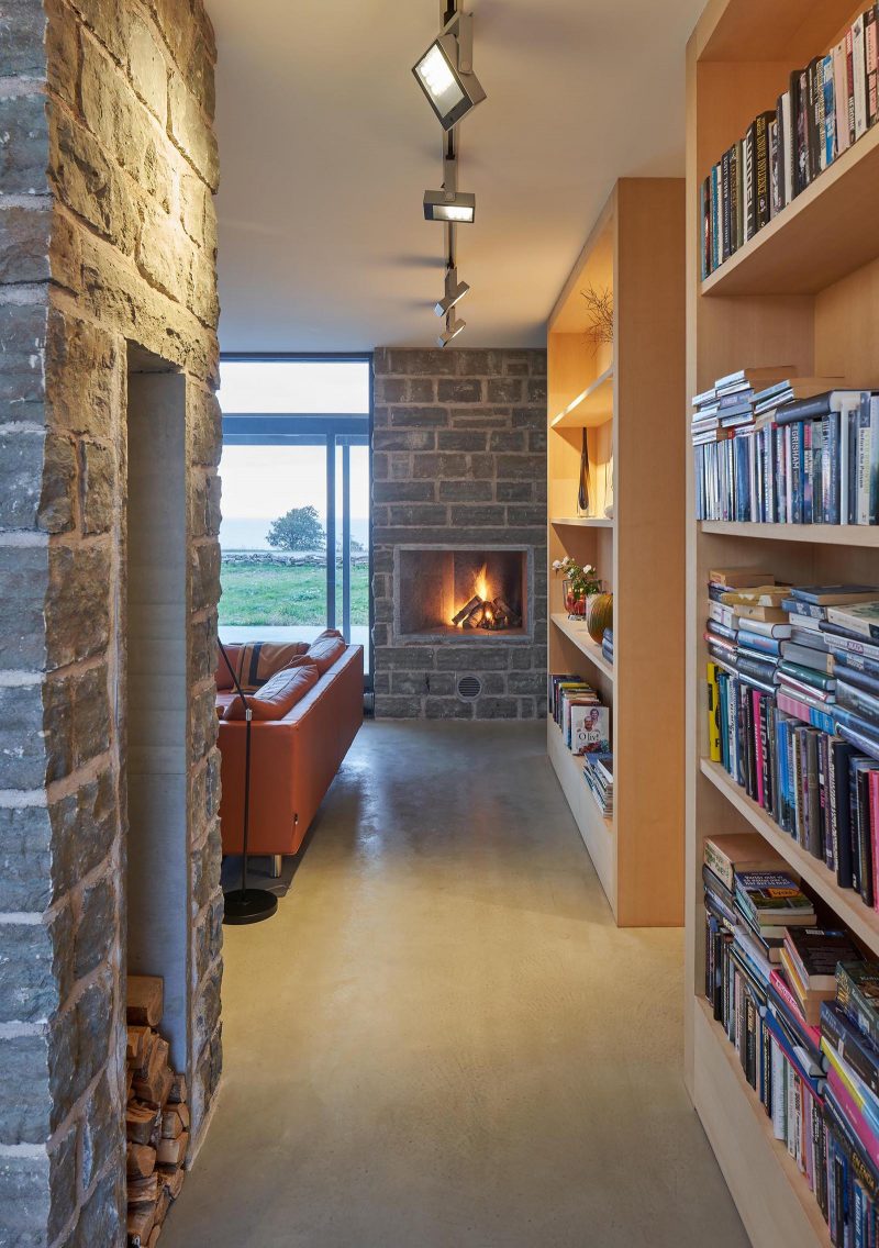 Современный летний дом со стенами из известняка, бетонным полом и деревянными книжными полками.