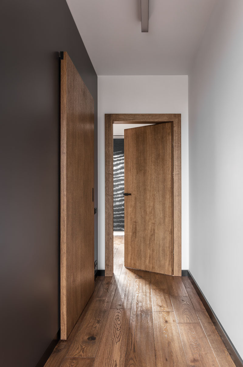 Современные апартаменты с деревянными дверями, темно-серыми акцентными стенами и деревянным полом.
