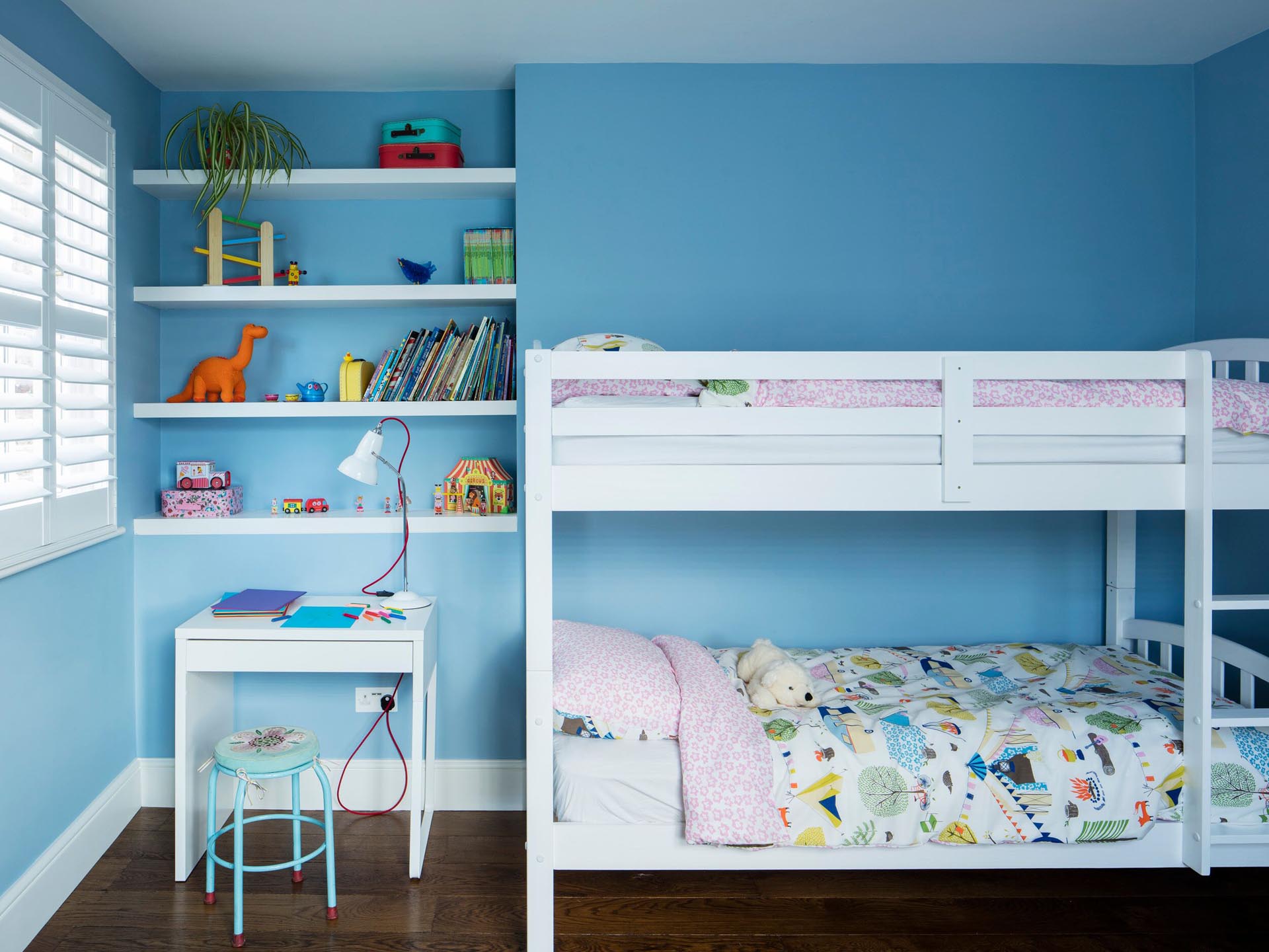В этой детской спальне ярко-синие стены создают красочный фон для белых двухъярусных кроватей, стеллажей и небольшого стола.