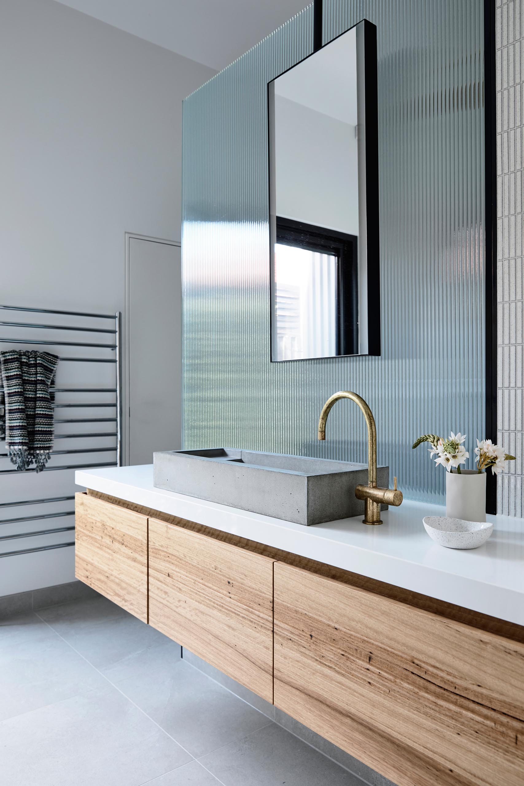 Современная ванная комната с длинным деревянным туалетным столиком, бетонной раковиной, бронзовым смесителем и текстурированной стеклянной панелью, скрывающей душ.