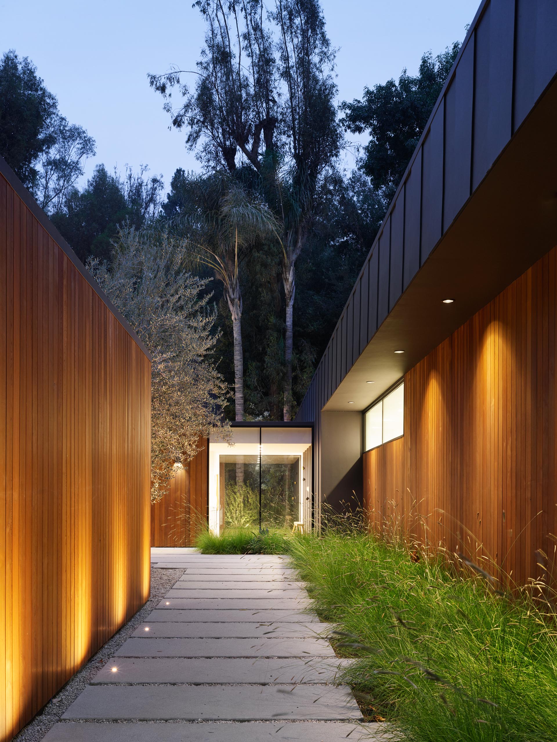 Дорожка из бетонной брусчатки, окаймленная дикой травой и освещением, ведет посетителей к входной двери.