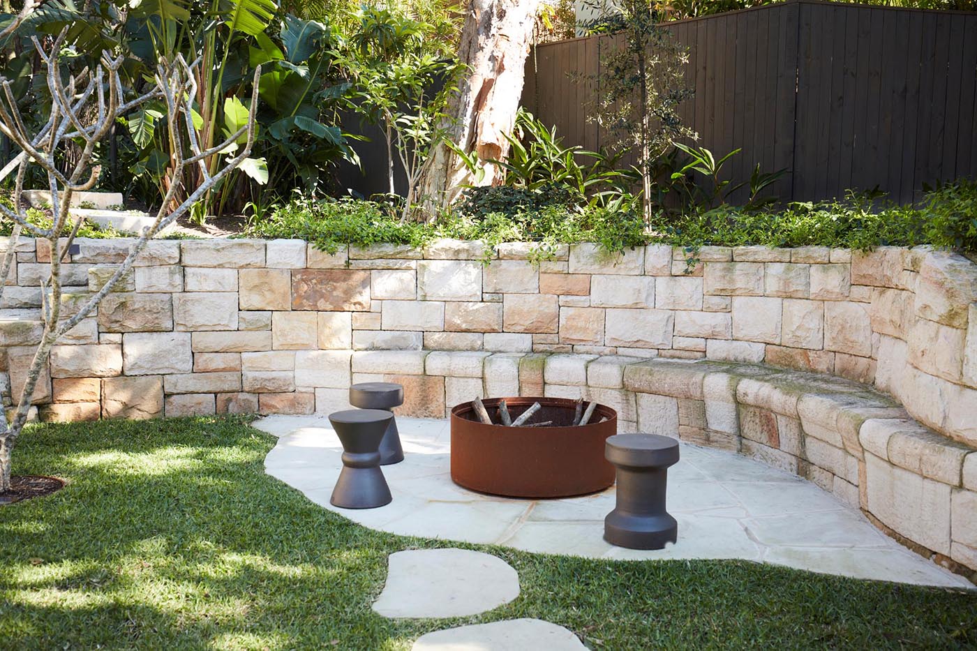 Стена из песчаника включает места для сидения вокруг костровой ямы и включает ступеньки, ведущие на верхний уровень сада.