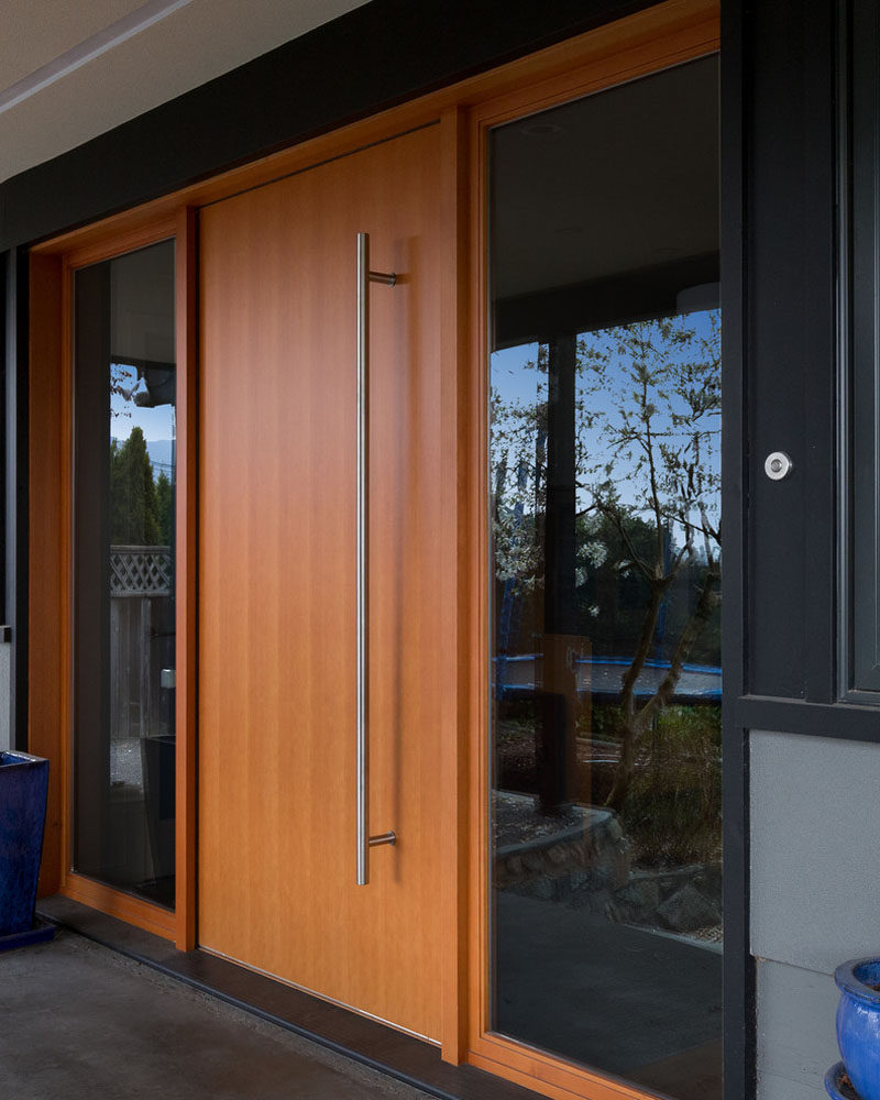 Эта дверь из светлого дерева имеет большую металлическую дверную ручку и обрамлена окнами с обеих сторон, что создает современный вход в этот семейный дом.