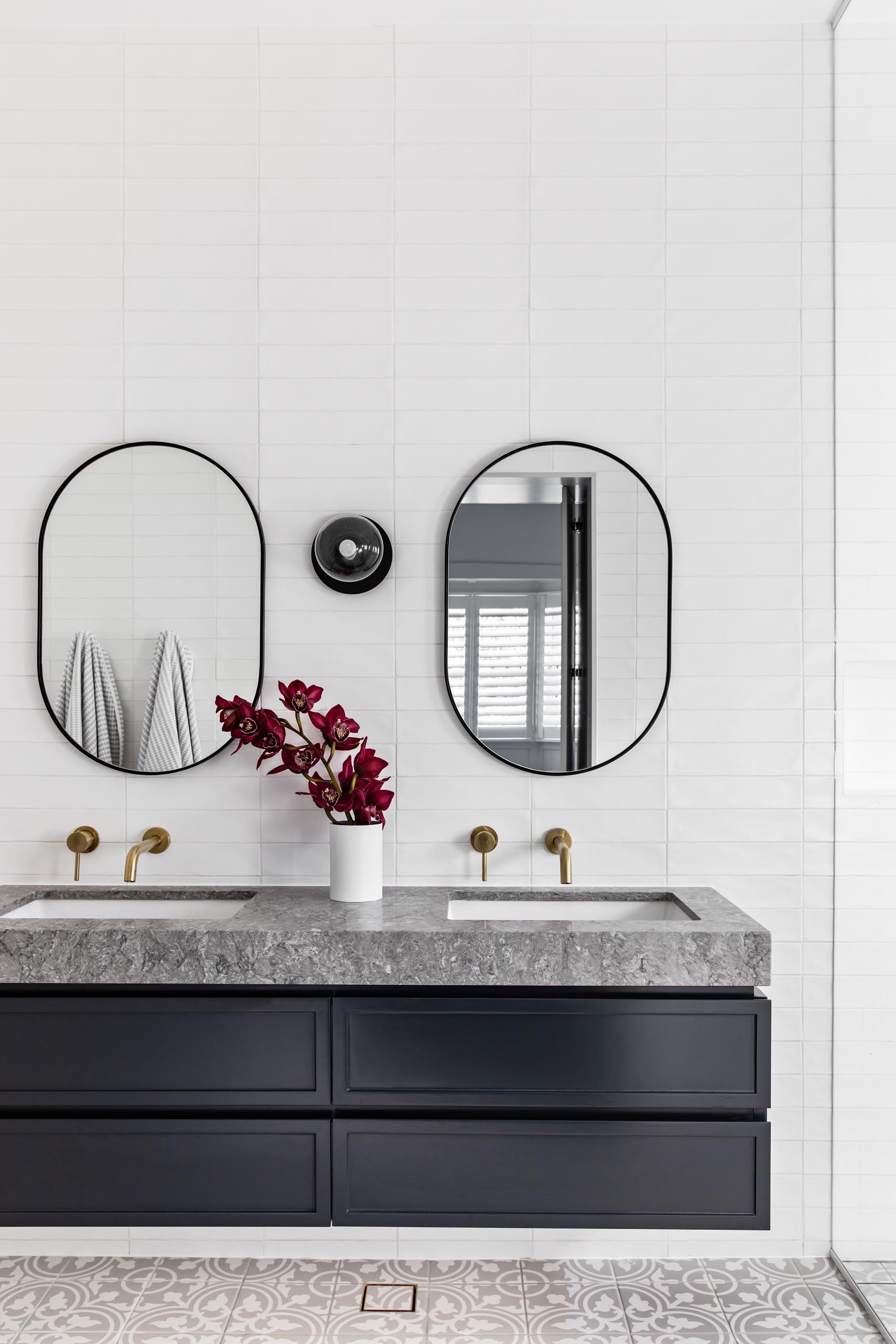 Современная ванная комната с узорчатой ​​плиткой на полу, плавающий черный туалетный столик с серым камнем, белая плитка метро, ​​покрывающая стены, овальные зеркала в черной рамке и латунная фурнитура.