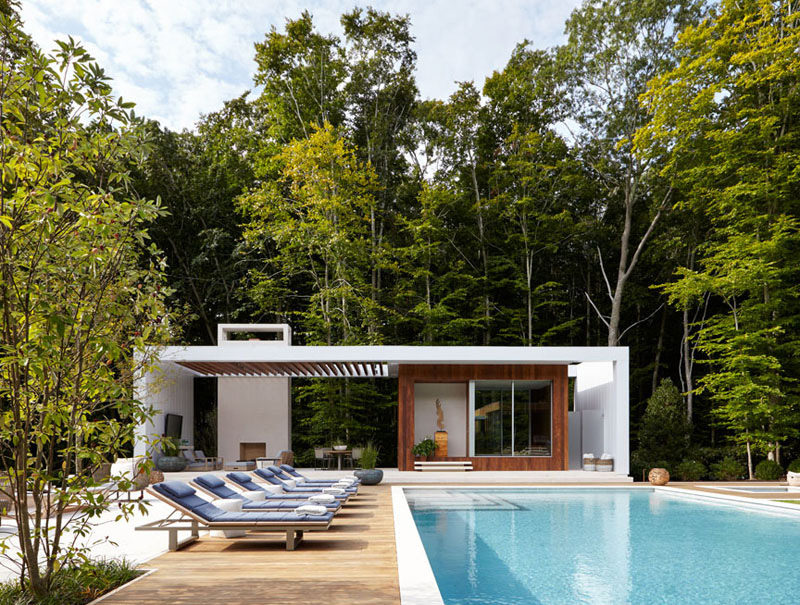 Бело-деревянный дом у бассейна с крытой открытой гостиной зоной с камином. Синие мягкие шезлонги расположены вдоль бассейна.