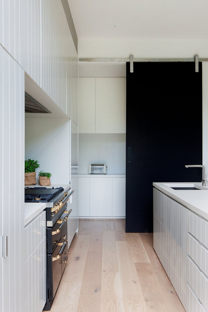 Черная раздвижная дверь сарая на этой современной кухне создает смелый контраст на фоне белой мебели и пола из светлого дерева.