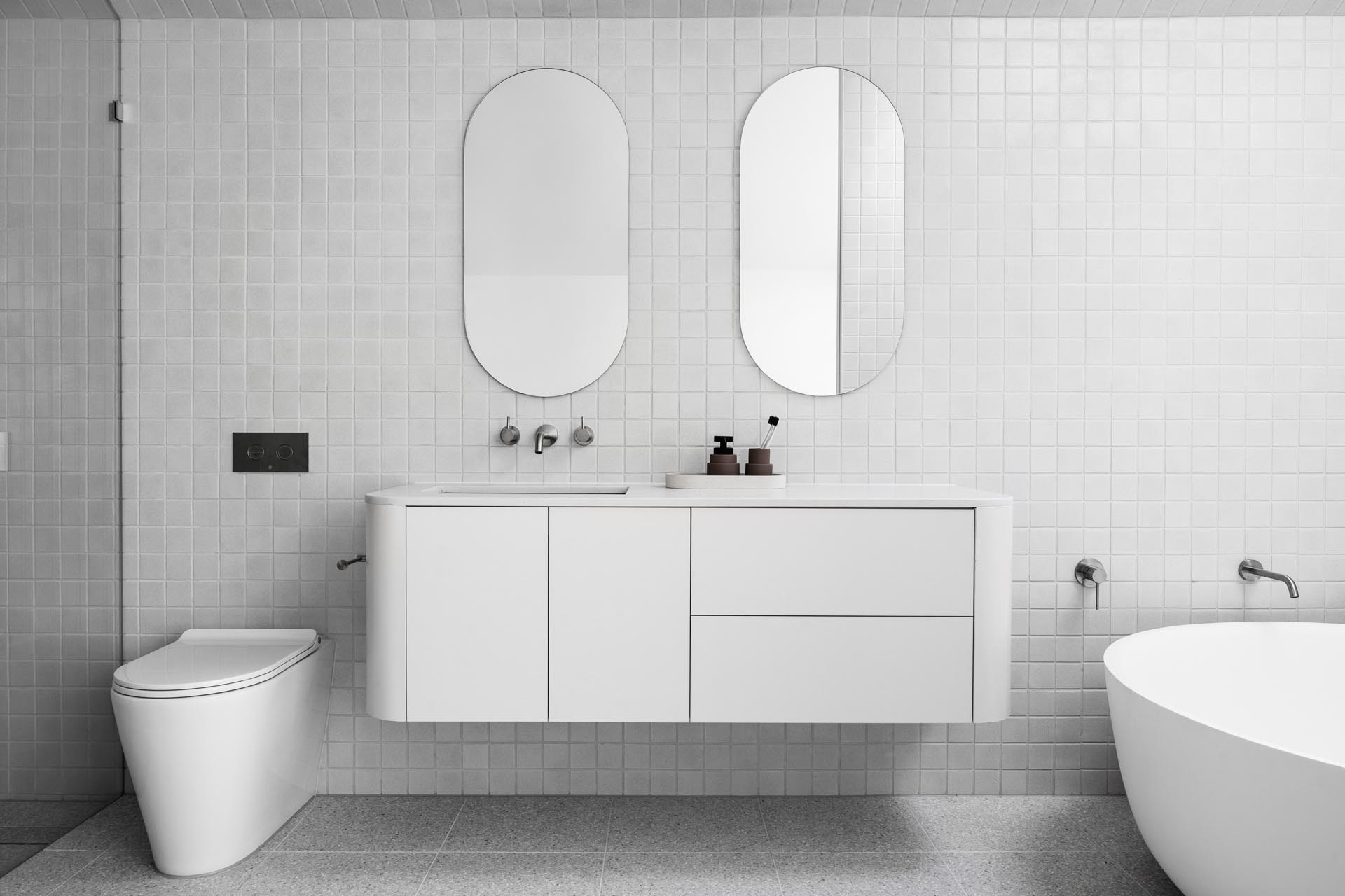 В этой современной ванной комнате квадратная серая плитка покрывает стены и пол, а зеркала в форме пилюль висят над плавающим белым туалетным столиком с изогнутыми углами. Один конец ванной комнаты отведен под душ, а в другом - круглая отдельно стоящая ванна, расположенная под потолочным окном.