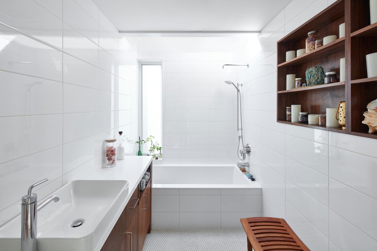 В этой современной ванной комнате белая плитка покрывает стены и переднюю часть встроенной ванны. Деревянный стеллаж на стене дополняет деревянный туалетный столик на противоположной стене.