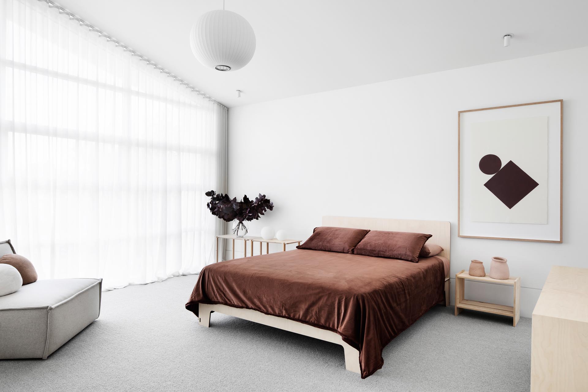 В этой главной спальне темно-коричневые постельные принадлежности контрастируют с более светлыми элементами в комнате, такими как пол, прикроватные тумбочки и стул с обивкой.