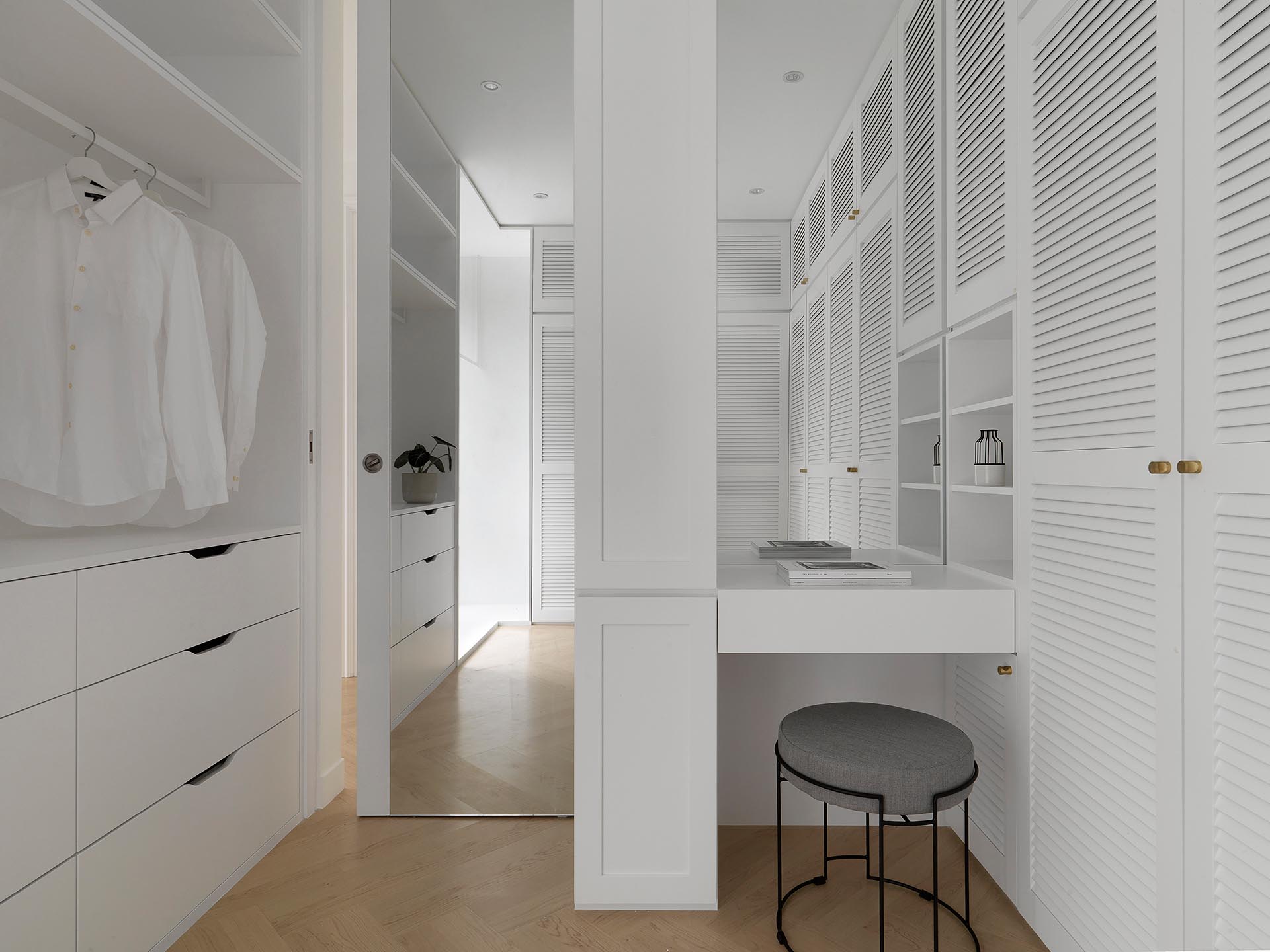 Современная гардеробная с белыми шкафчиками, маленьким туалетным столиком и зеркалом от пола до потолка за дверью.