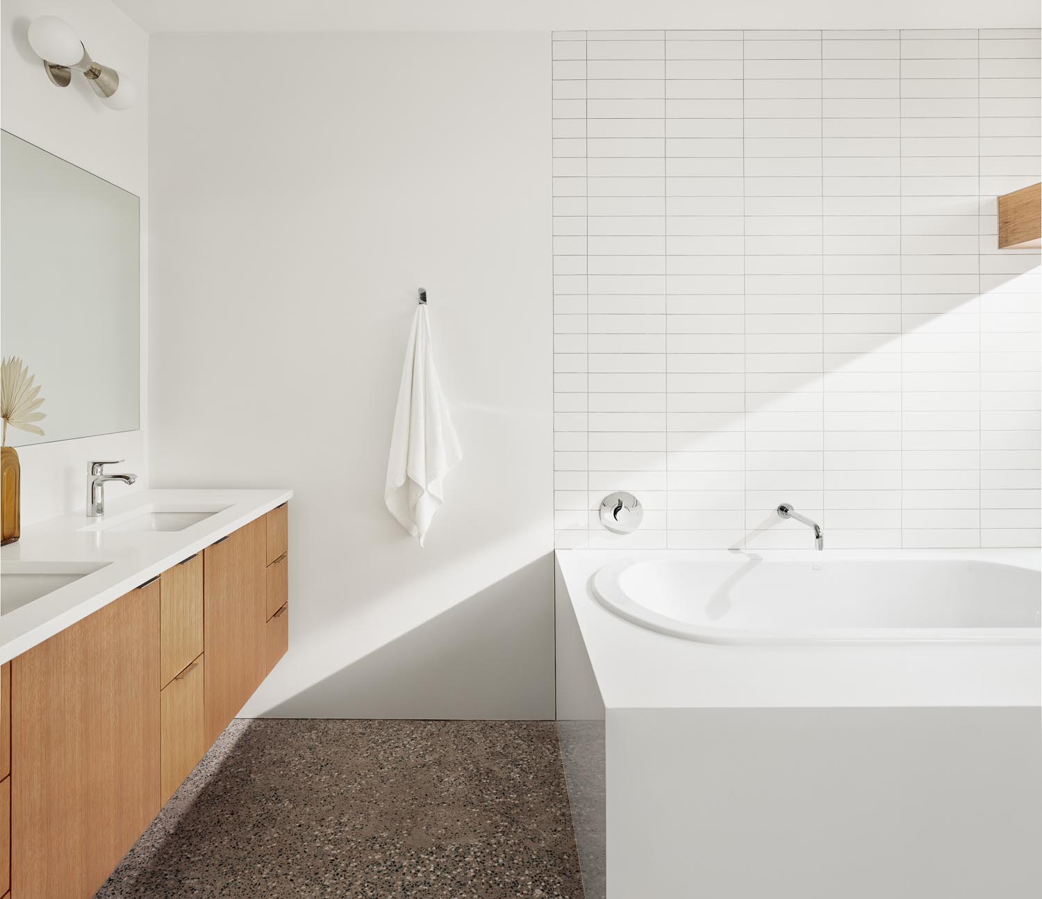 В этой современной ванной комнате есть парящий деревянный туалетный столик, встроенная ванна и стены, облицованные белой прямоугольной плиткой.